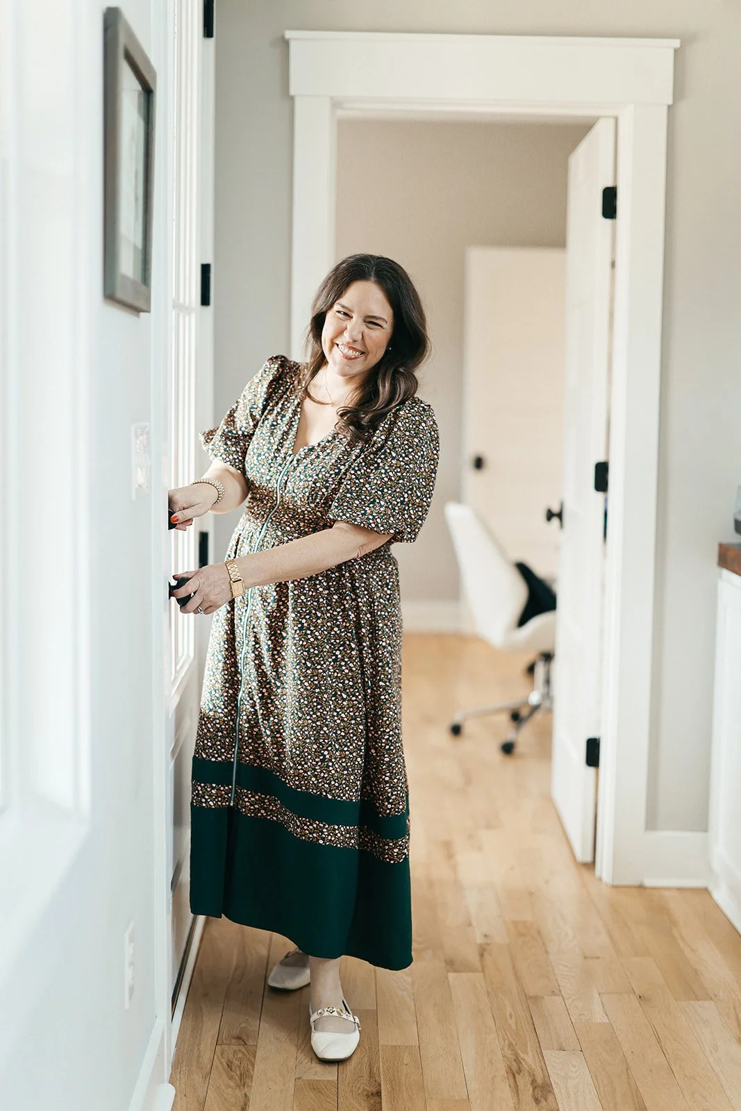 Woman in a floral dress smiling and closing a cabinet door in a home interior with hardwood floors.
