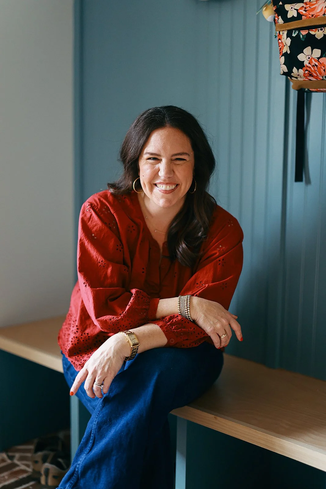 A woman with dark wavy hair, smiling, wearing a red blouse with eyelet details, dark blue jeans, hoop earrings, and a gold watch, sitting on a wooden bench against a blue wall.