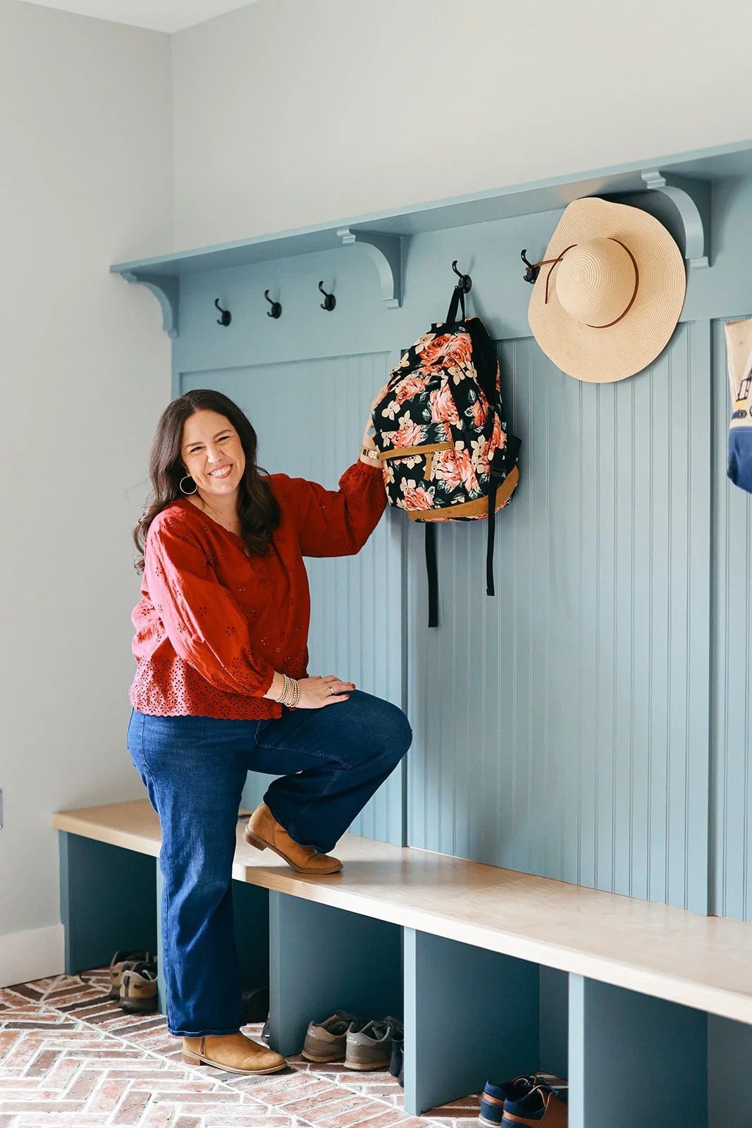 A woman in a red blouse and blue jeans smiling and holding a floral backpack in a hallway with a blue shelf and hooks, a beige bench, and shoes underneath.