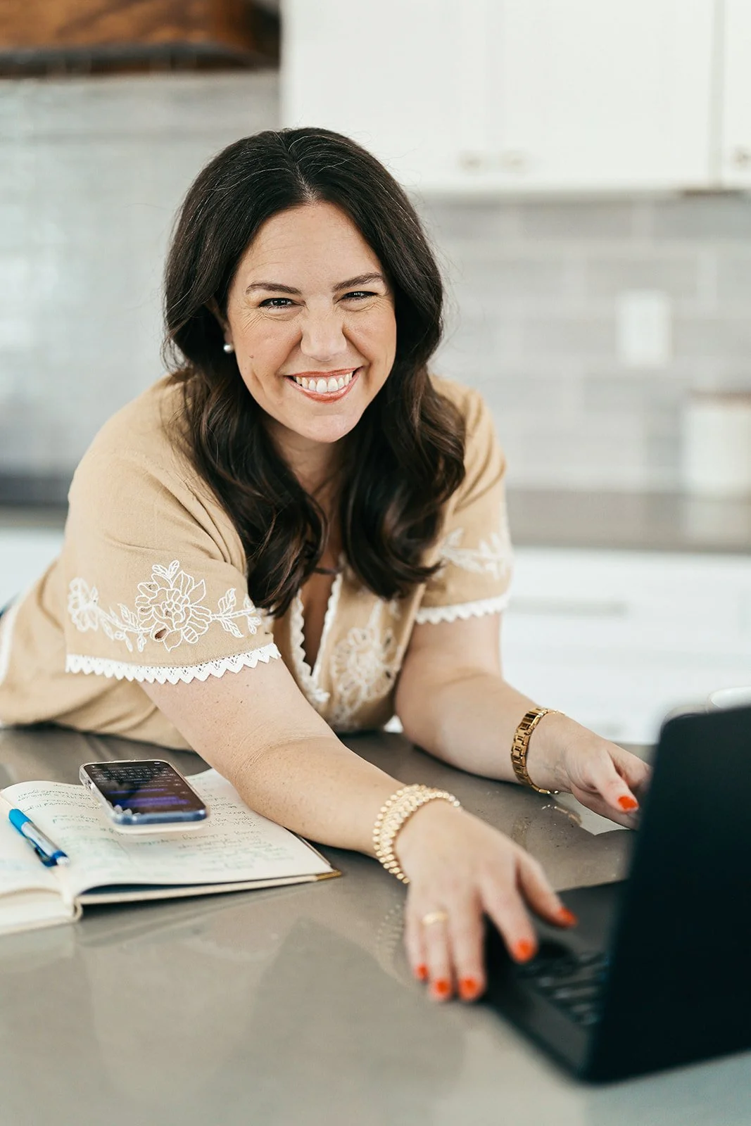 A woman smiling at a kitchen counter with a laptop, notebook, pen, and smartphone, wearing a beige embroidered top and jewelry.