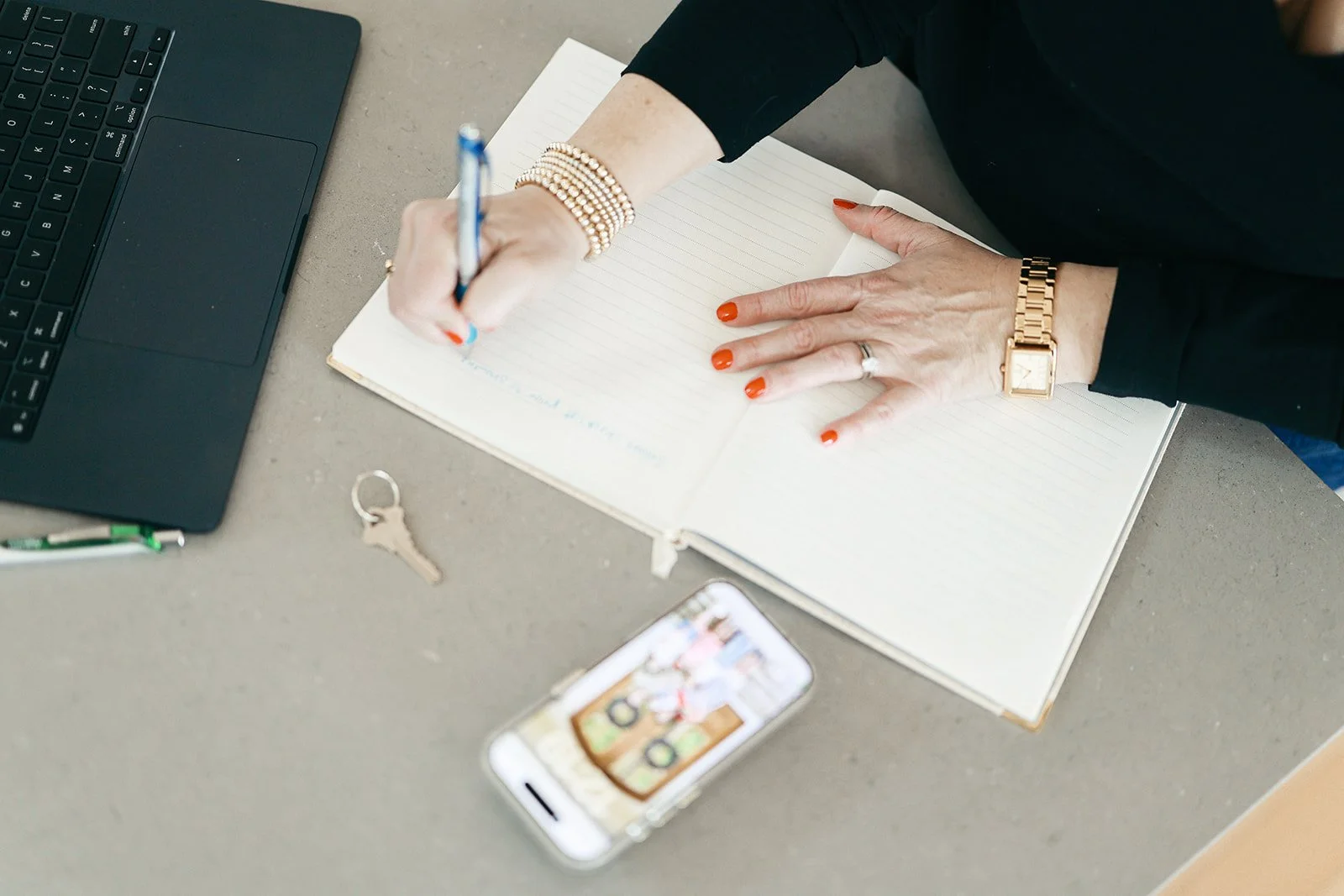 A person writing in a notebook at a desk with a laptop, keys, a pen, and a smartphone showing a photo of someone in front of a wooden background. The person has orange nail polish, a bracelet, and a gold watch.