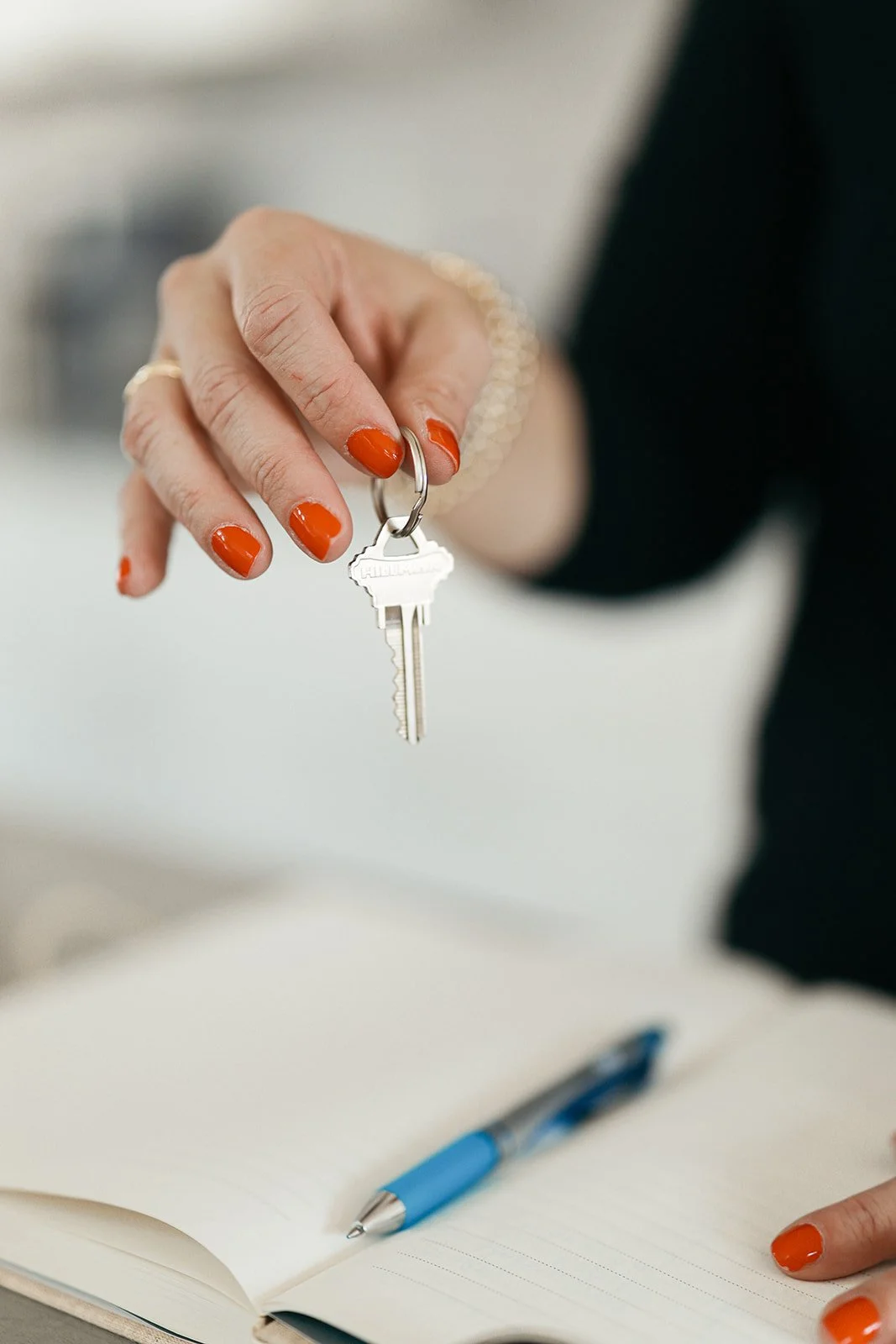 A woman with red nail polish and wearing jewelry holds a key ring with a key, over an open notebook with a blue pen.