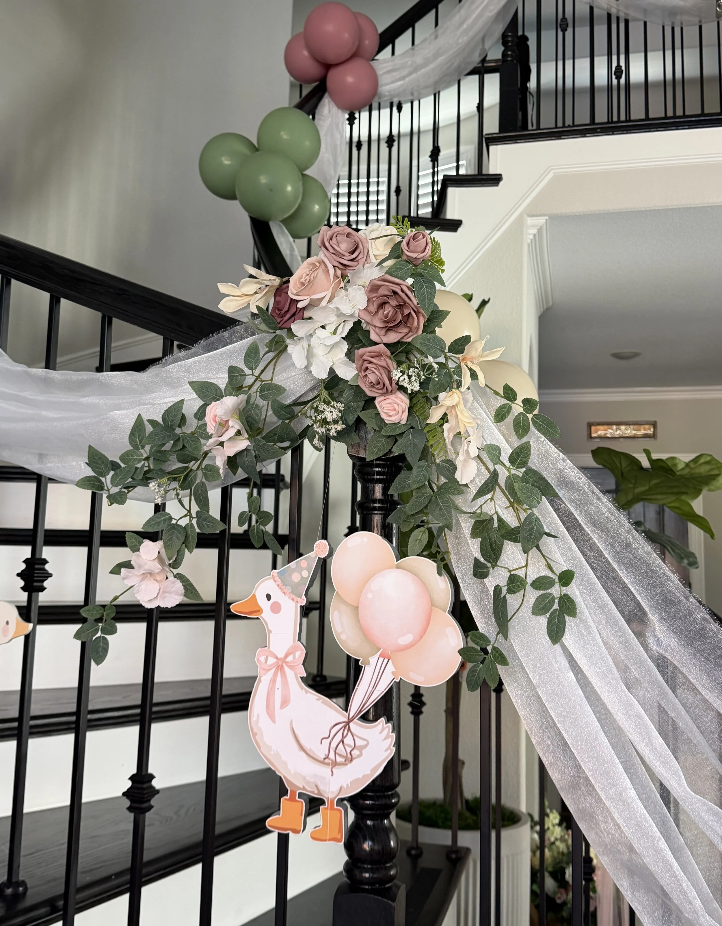 Decorated staircase with pink, green, and beige balloons, a paper cutout of a smiling goose for a one silly goose theme, surrounded by pink roses, white flowers, and green leaves, with white sheer fabric draped along the  railing in Frisco Texas home