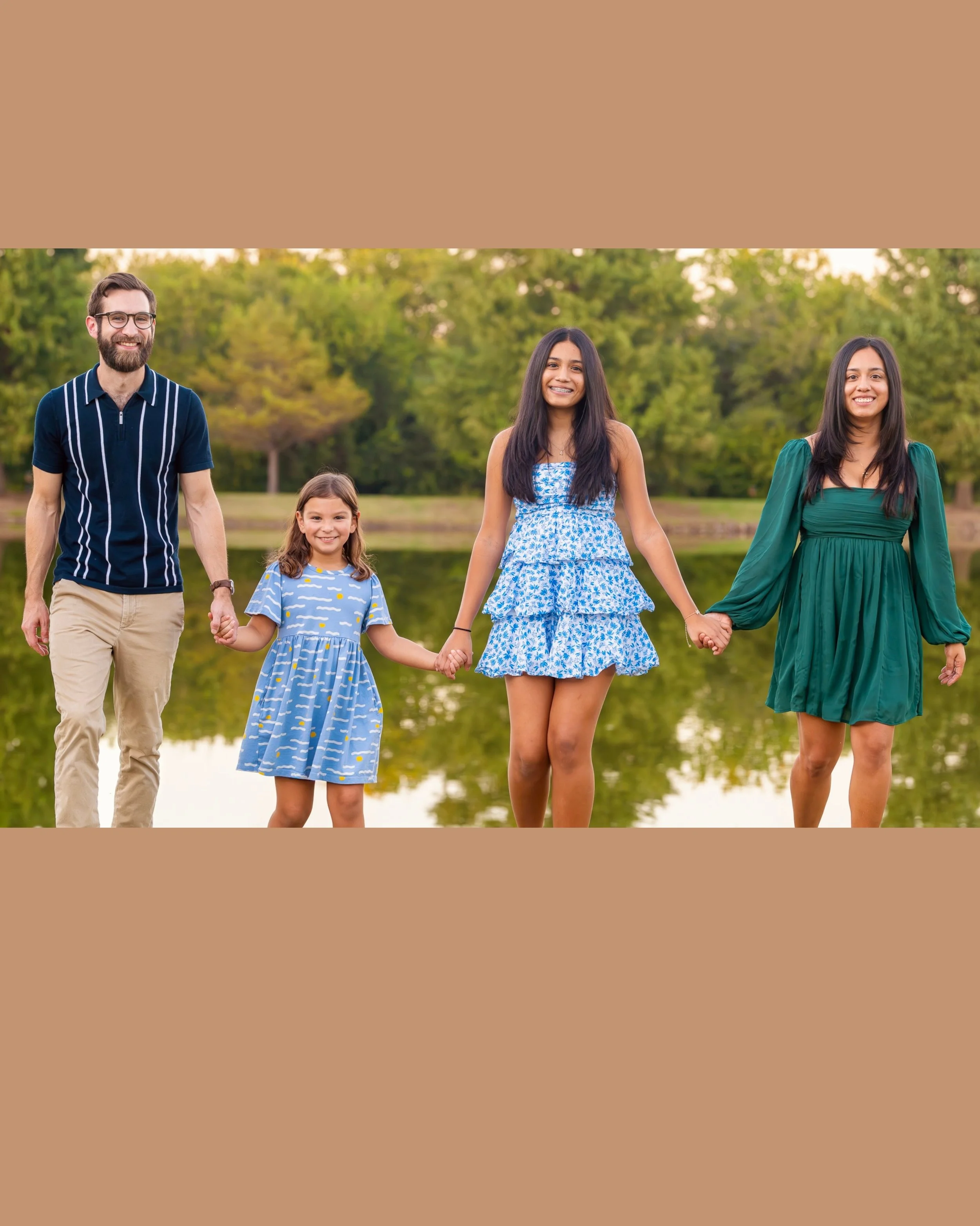 A family of four, including a man, woman, teenage girl, and young girl, holding hands and walking outdoors near a pond with trees in the background on a sunny day.
