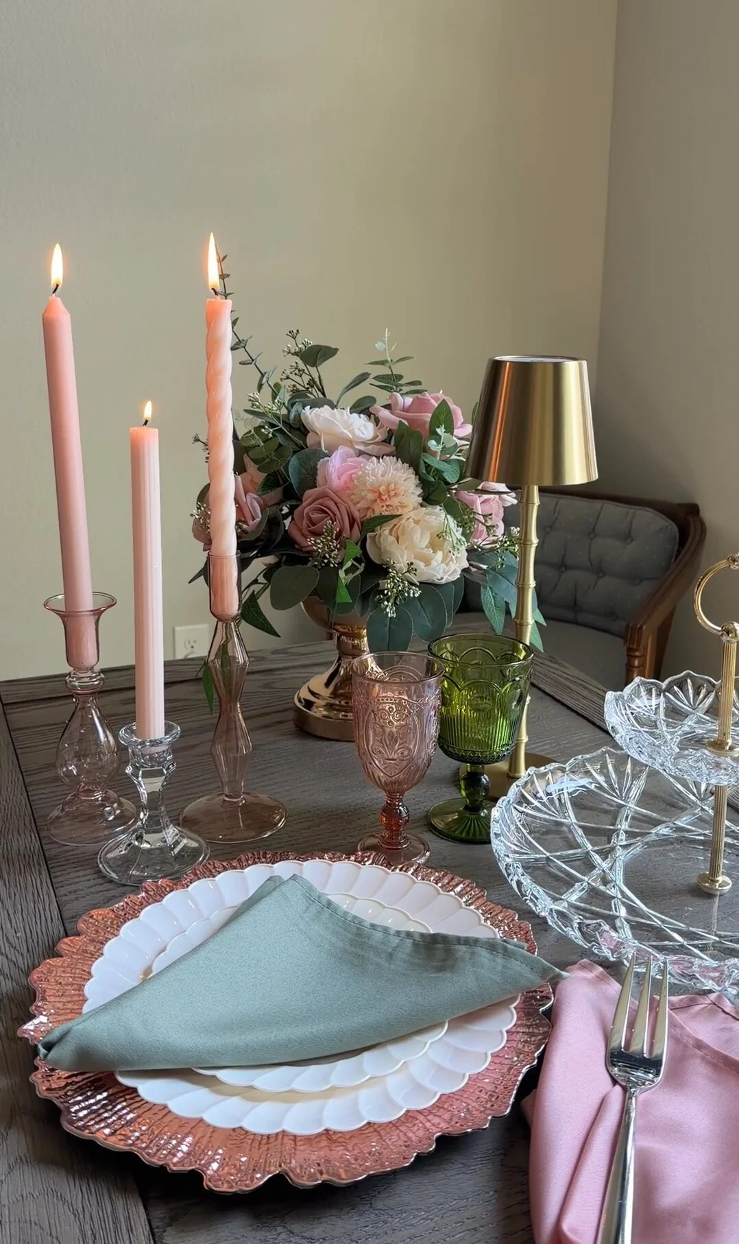 A table decorated for a special occasion with pink candles, a floral centerpiece with roses and greenery, colorful glassware, elegant plates with napkins, and a brass table lamp, set against a neutral wall and a wooden side chair.