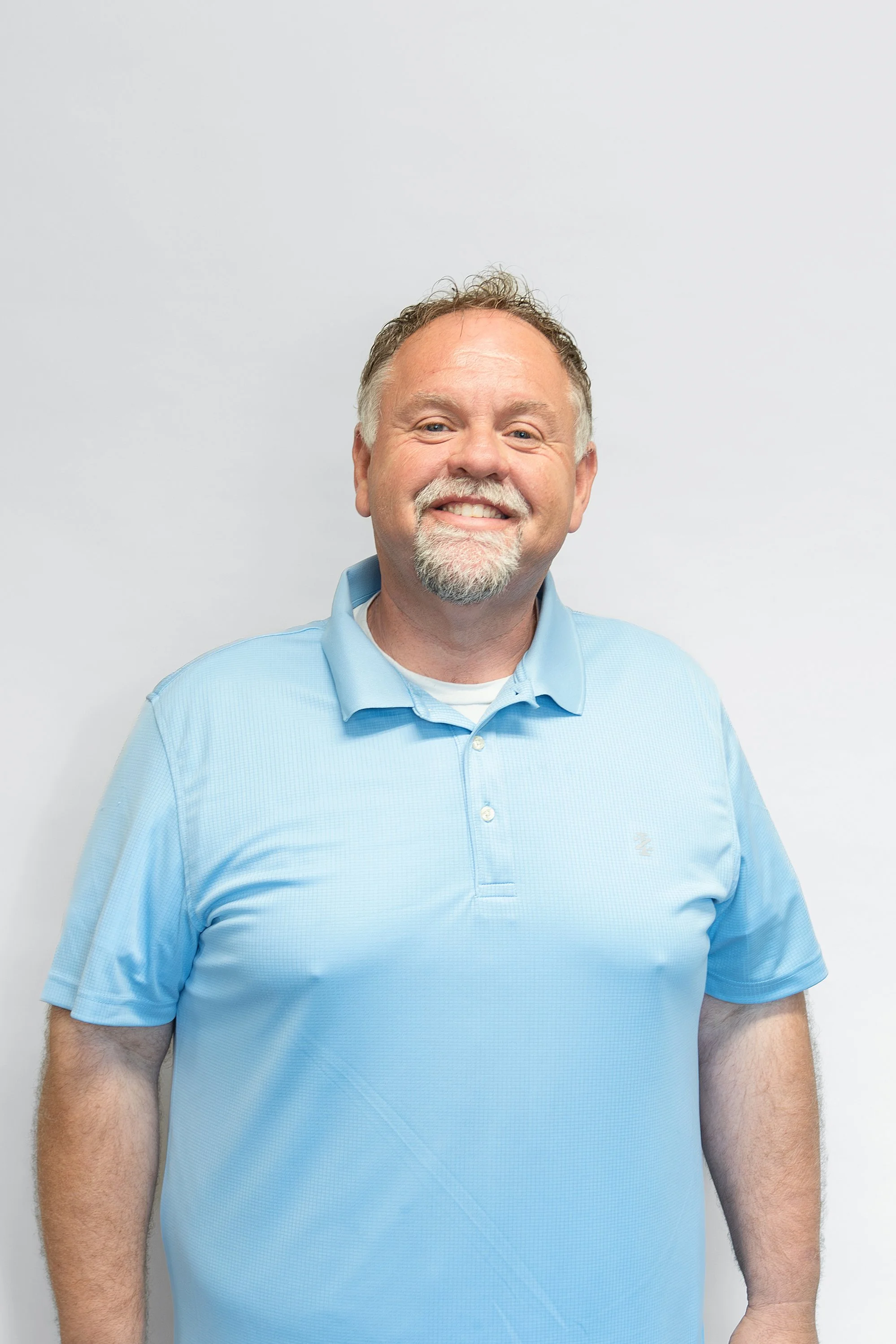 A middle-aged man with a beard and mustache, smiling, wearing a light blue collared shirt, standing against a plain white background.