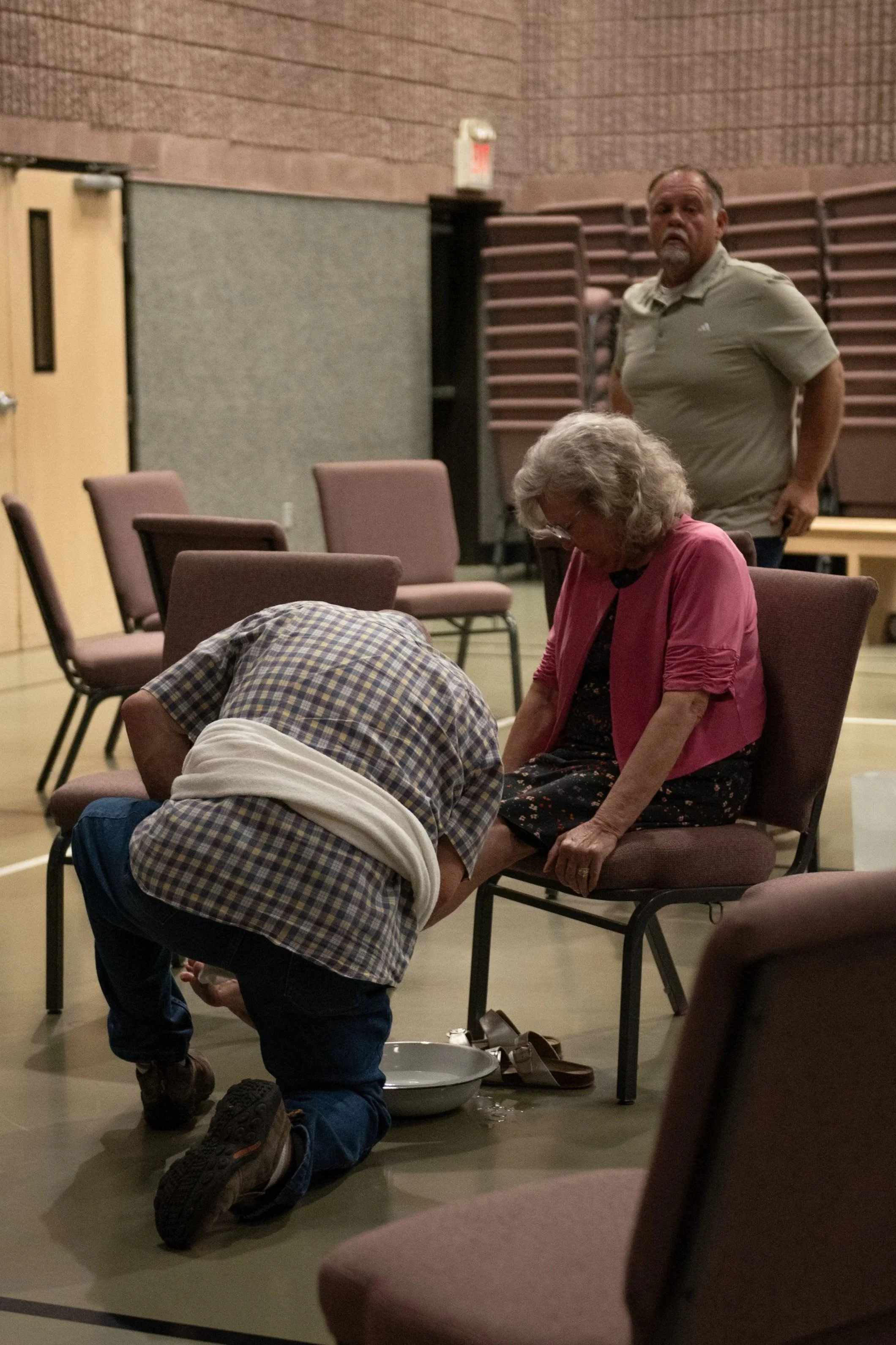 A man and a woman seated on chairs in a large room with stacked chairs in the background. The man is standing, looking towards the camera, and the woman appears to be praying with her head bowed and hands on her lap. The floor has some items, including shoes and a basin.
