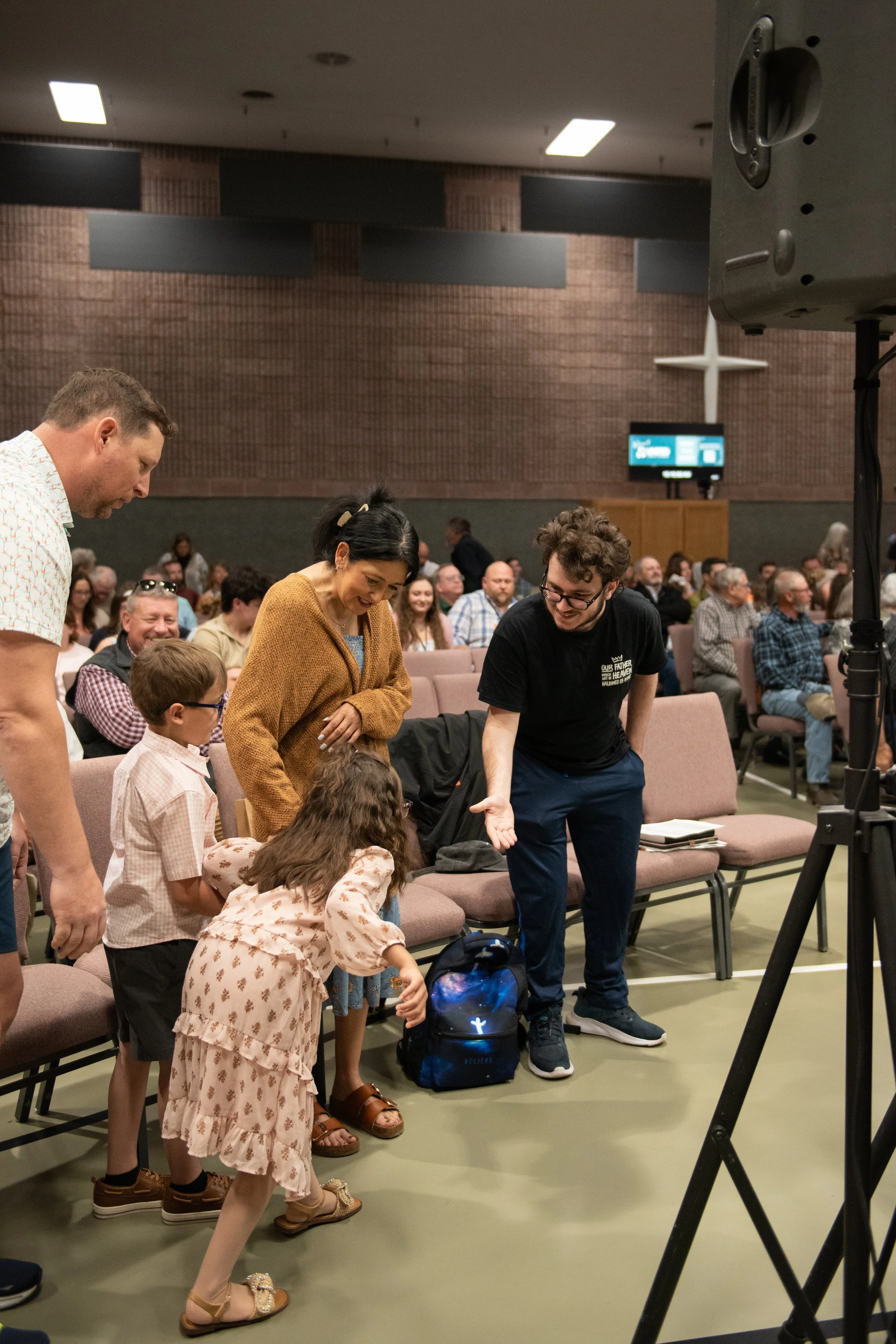 People gather in a church or community center, some sitting and some standing, engaged in a group activity or discussion. The room has brick walls, ceiling lights, and a large cross at the back.