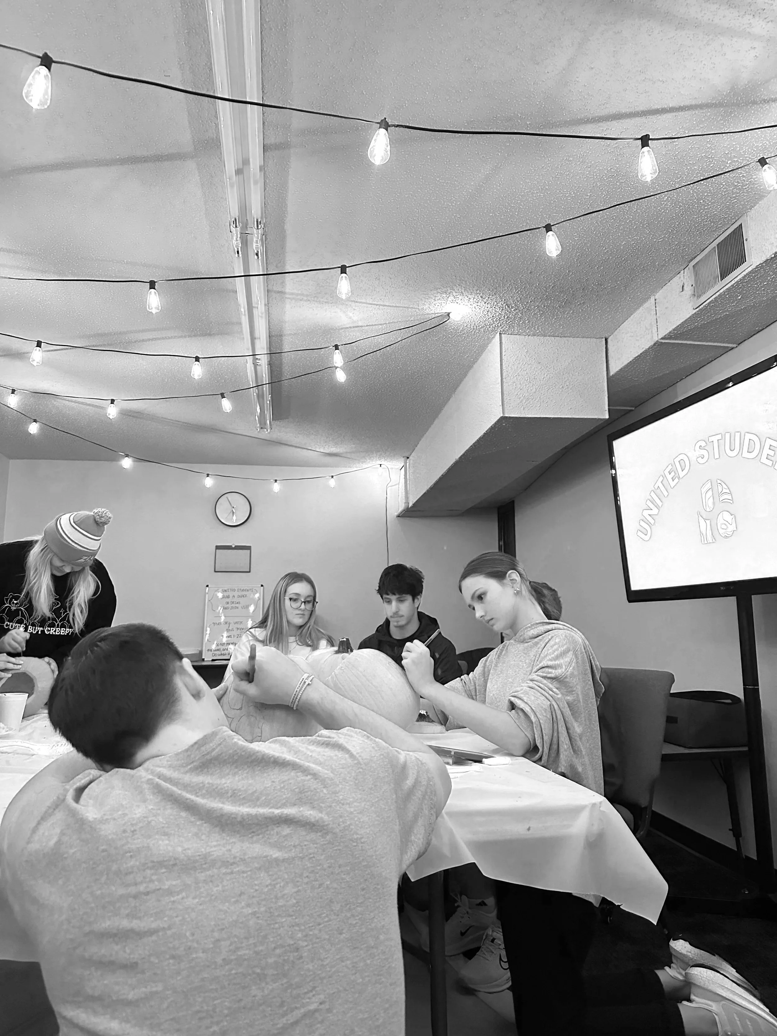 A group of young people sitting around a table, with some people getting tattooed by others in an indoor setting decorated with string lights and a wall clock.