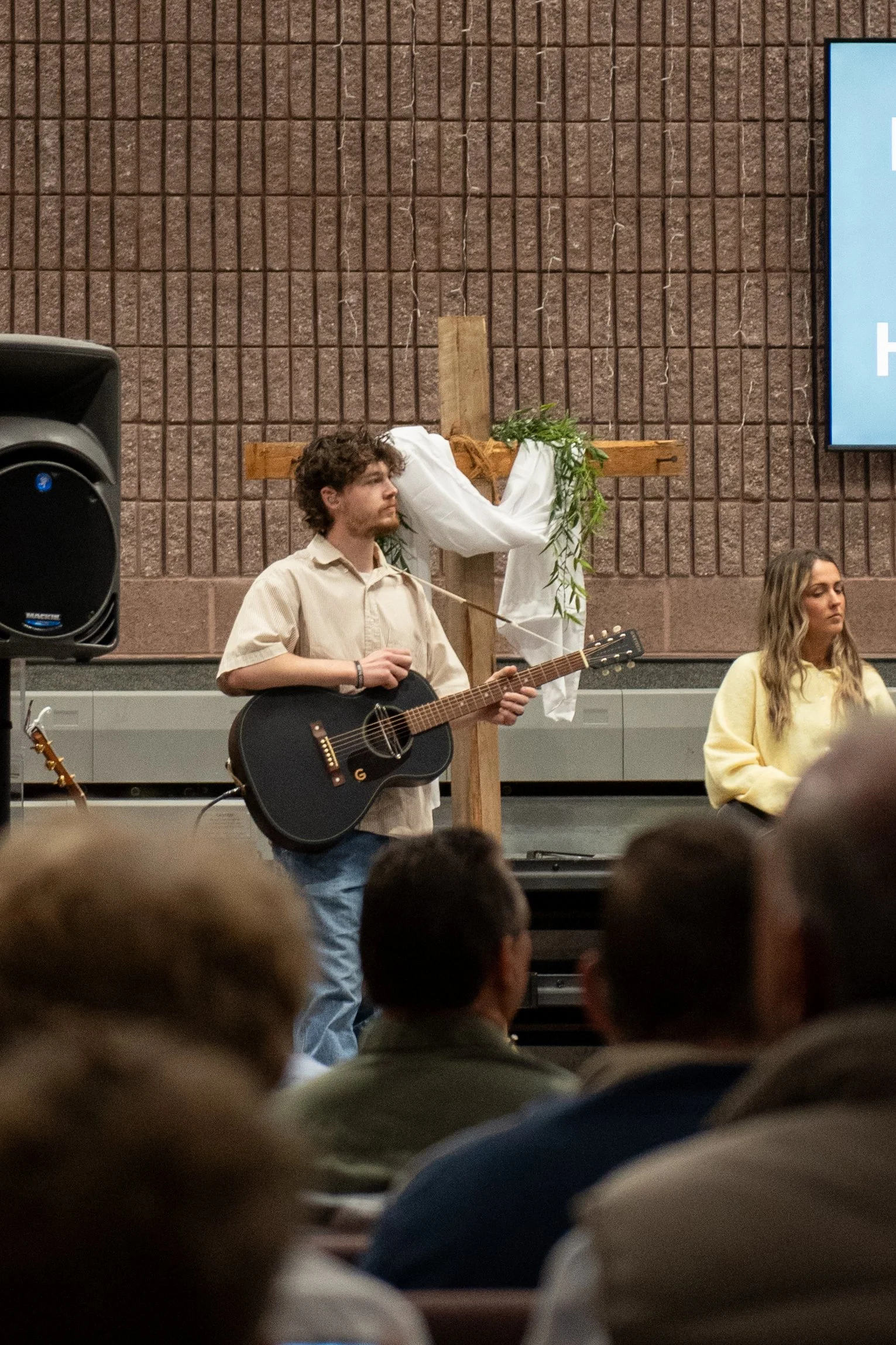 A young man with curly hair and a beard playing a black acoustic guitar during a church service or concert, standing in front of a large wooden cross decorated with greenery and white cloth, with a congregation visible in the foreground.