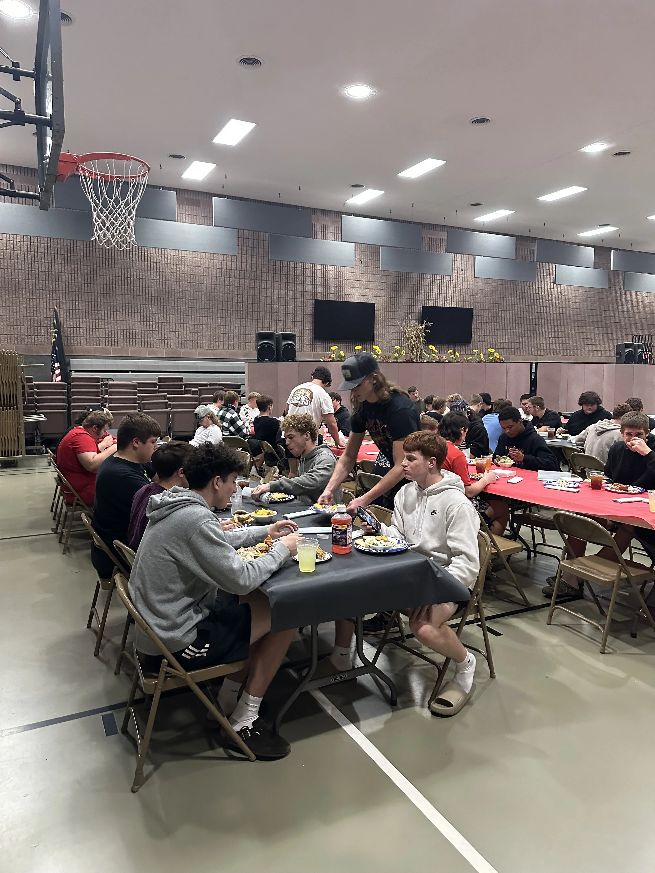Group of teenagers sitting at long tables in a gymnasium, eating food, with some standing and serving.