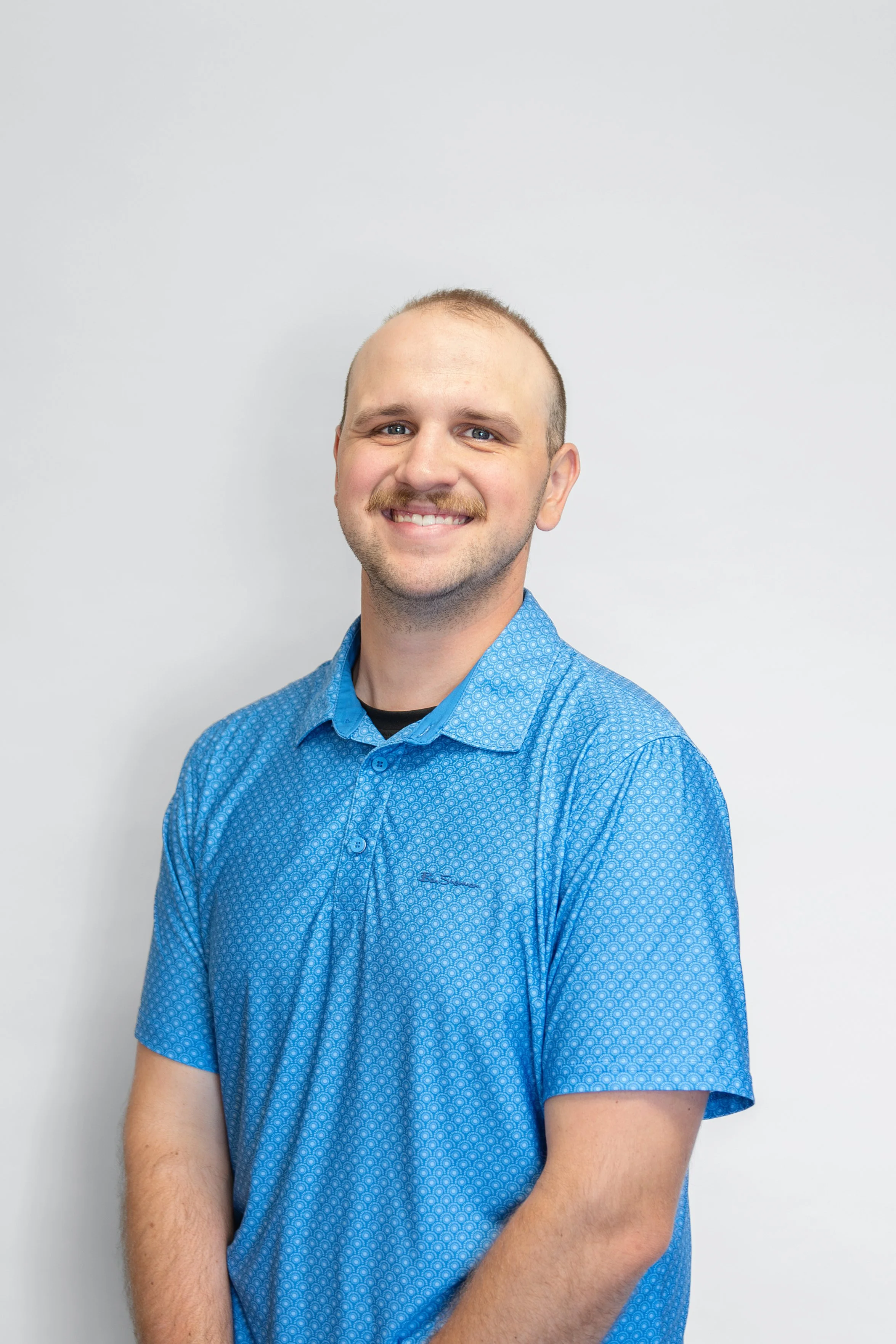 A smiling man with a mustache and short hair wearing a blue patterned short-sleeve shirt, standing against a plain white background.