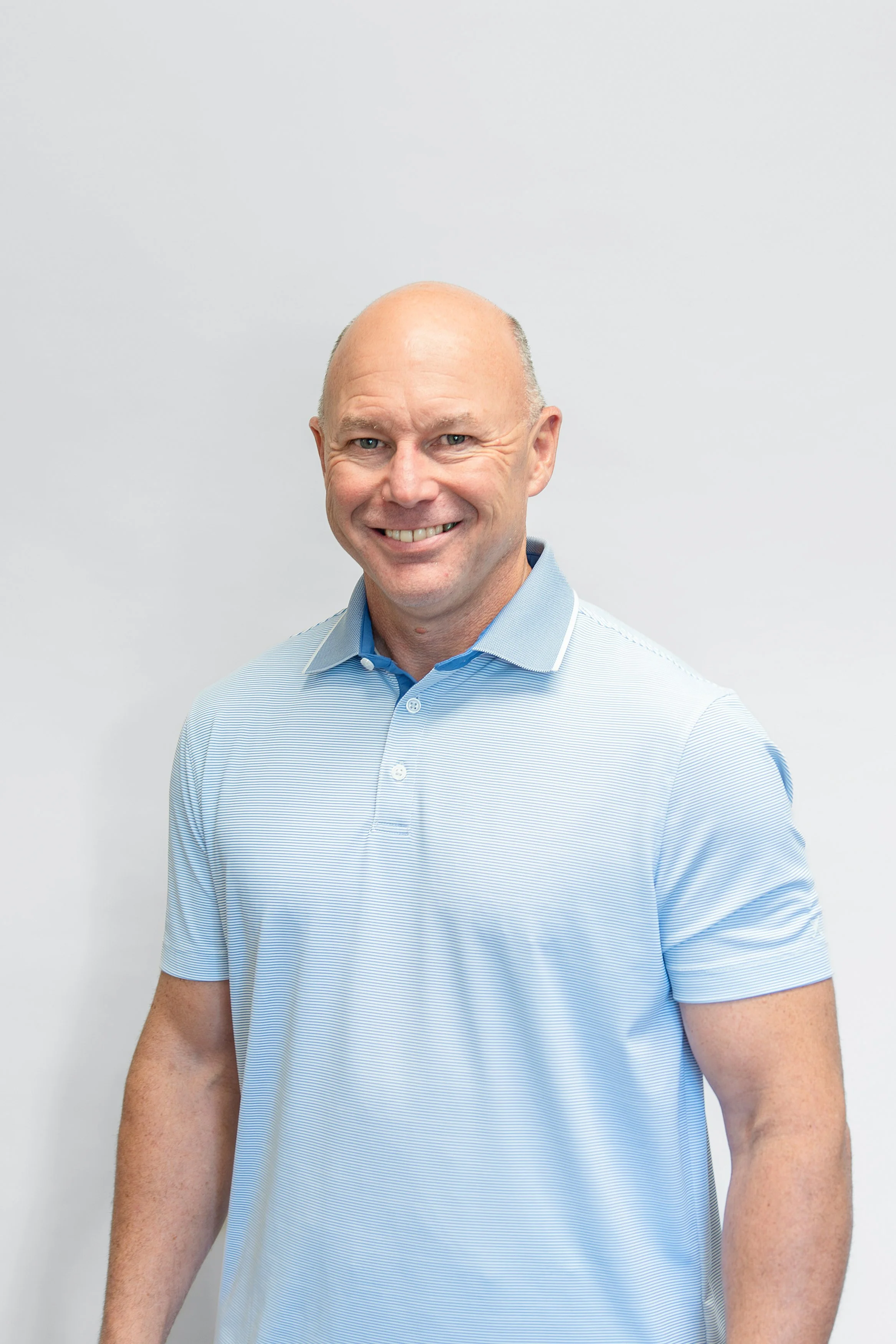 A smiling middle-aged man with a bald head wearing a light blue short-sleeve button-up shirt, standing against a plain white background.