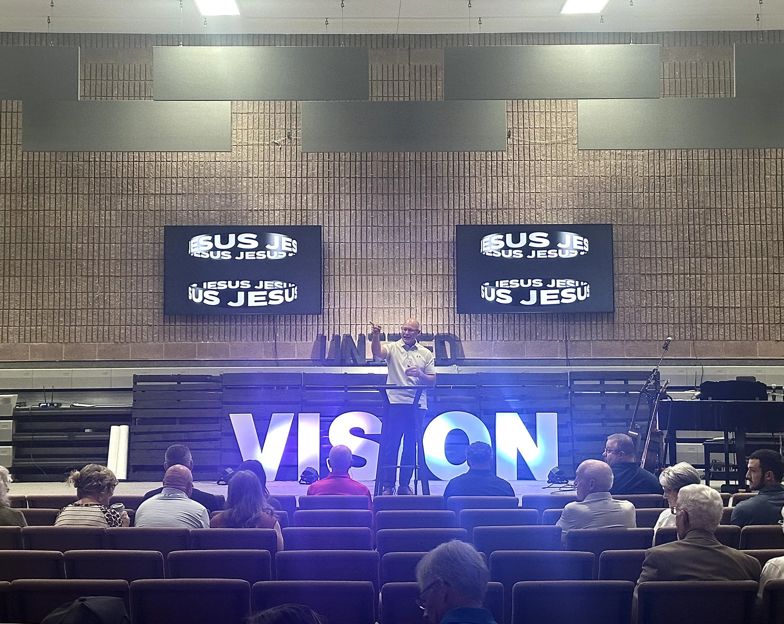 A man speaking at a church service, with a large illuminated sign that says 'VISON' behind him. The setting appears to be a church or auditorium with brick walls and multiple screens displaying the word 'JESUS'.