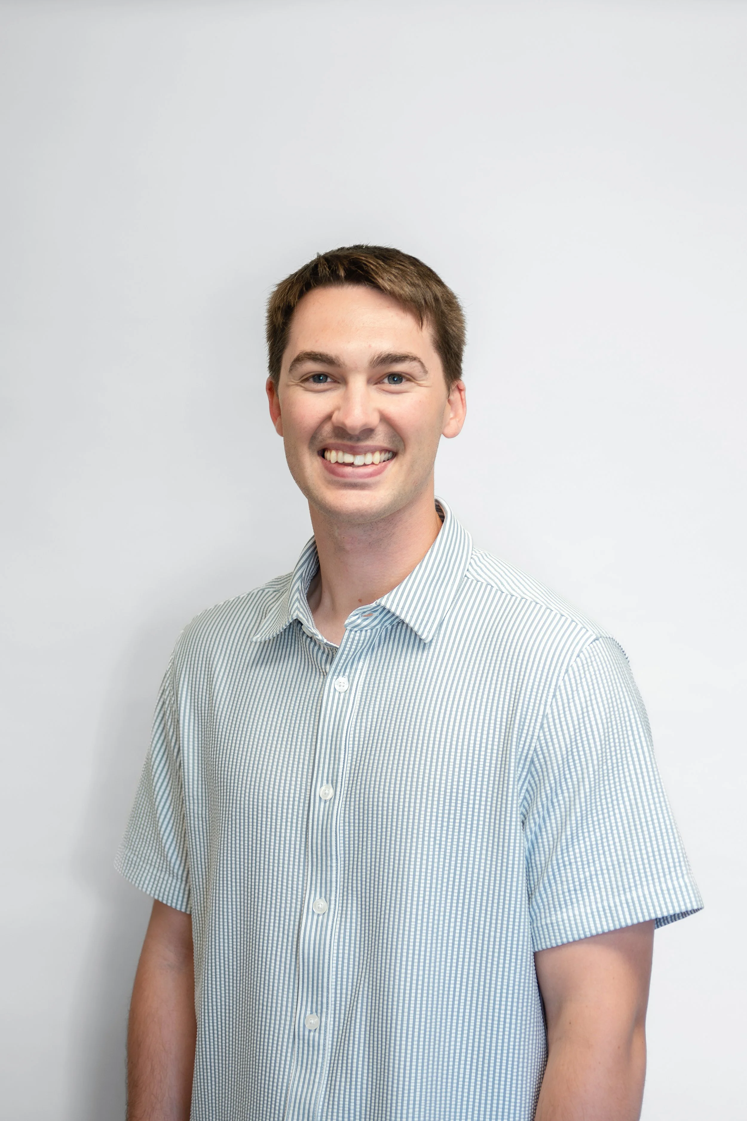 A young man with short brown hair, blue eyes, and a big smile, wearing a light blue and white striped button-up shirt, standing against a plain light gray background.