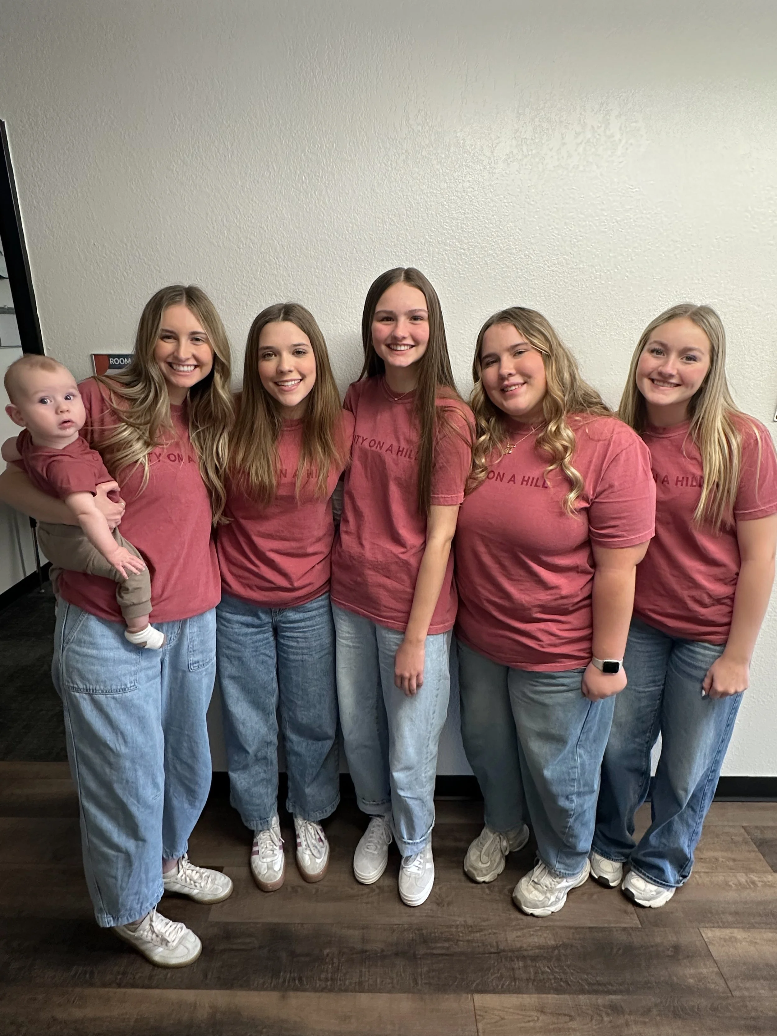 Group of five young women and one young girl standing together against a plain wall, all wearing matching maroon t-shirts and light blue jeans.