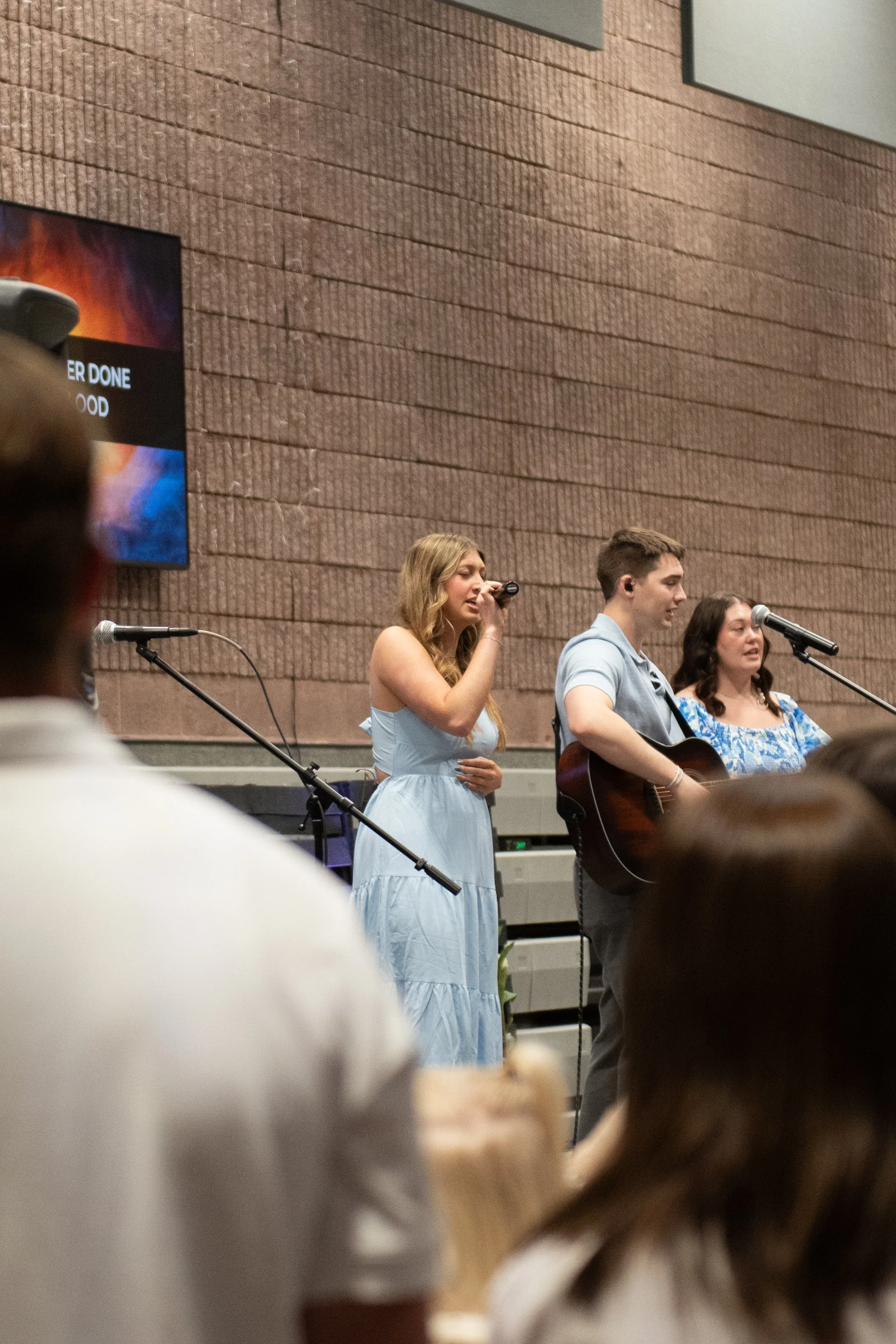 Young woman in a light blue dress singing into a microphone, accompanied by a young man playing guitar and another young woman singing, on a stage with a brick wall background, audience in foreground.