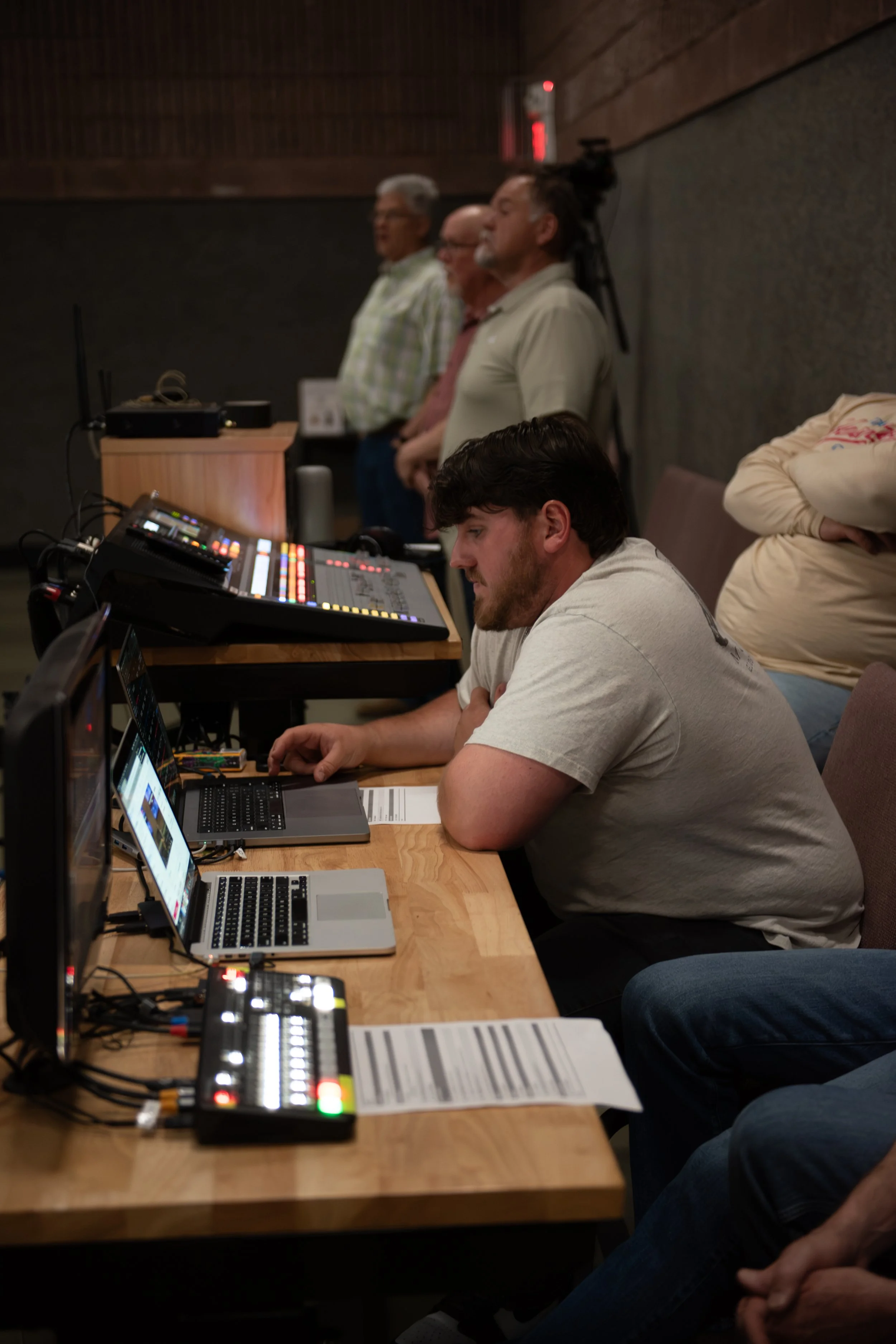People in a sound studio, some standing and some seated at audio equipment, including laptops and audio mixers.
