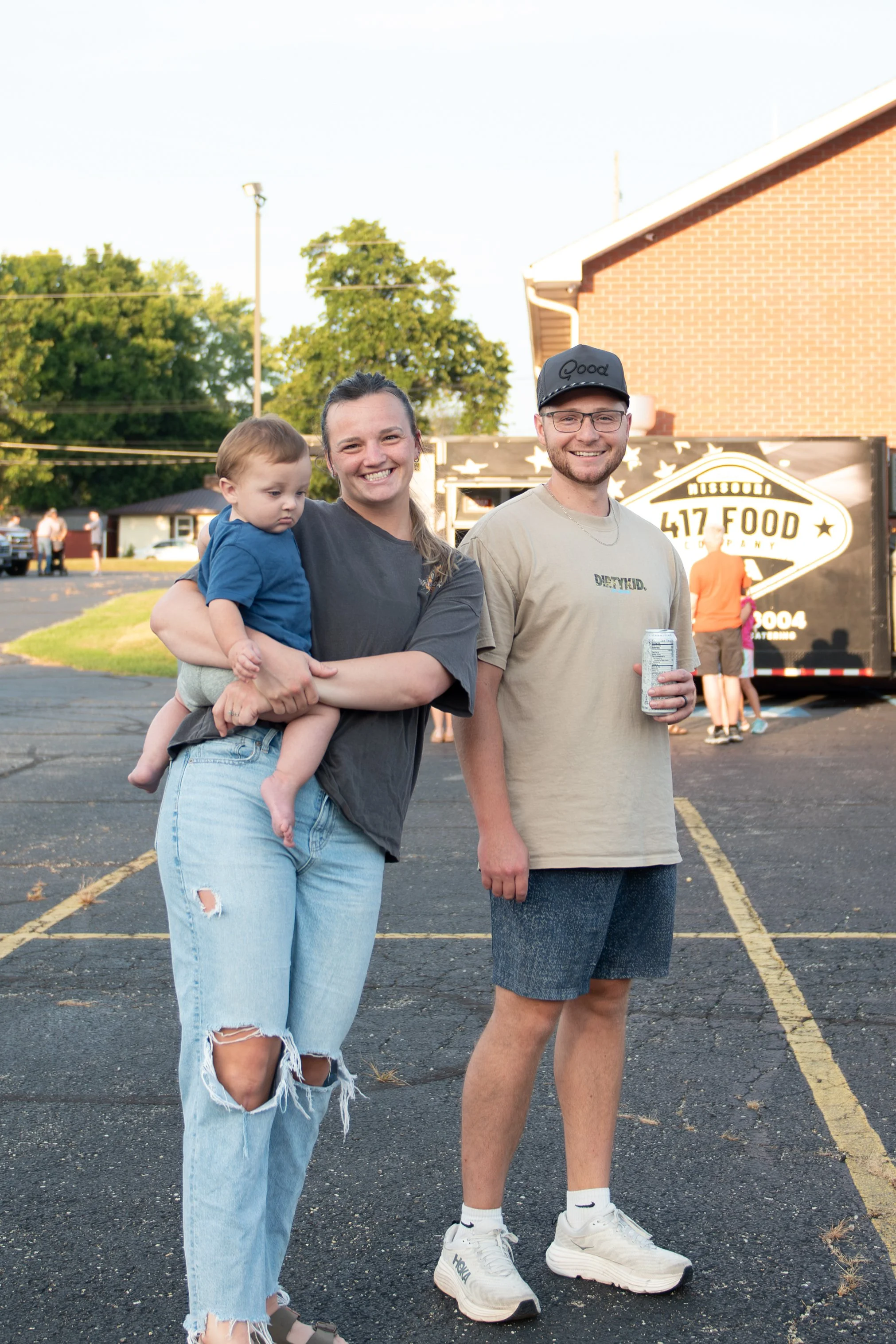 A group of three people standing outdoors in a parking lot, smiling at the camera, with trees, a brick building, and a food truck in the background. One adult woman is holding a child, and a smiling man with glasses and a cap is standing beside them, holding a beverage can.
