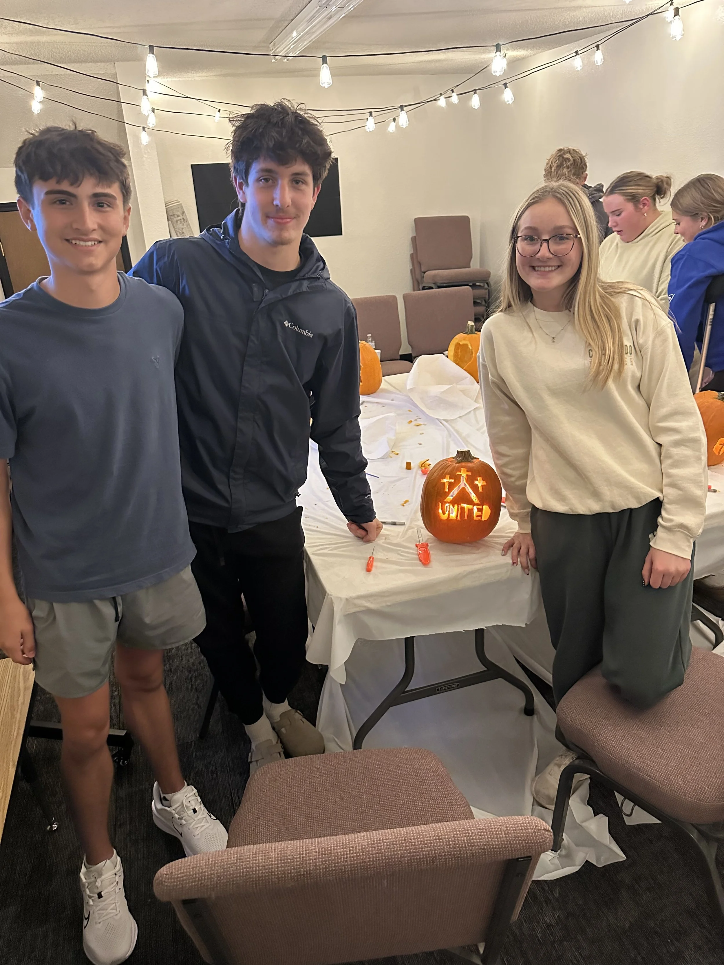 Four young people standing around a table with carved pumpkins with lights and people in the background.