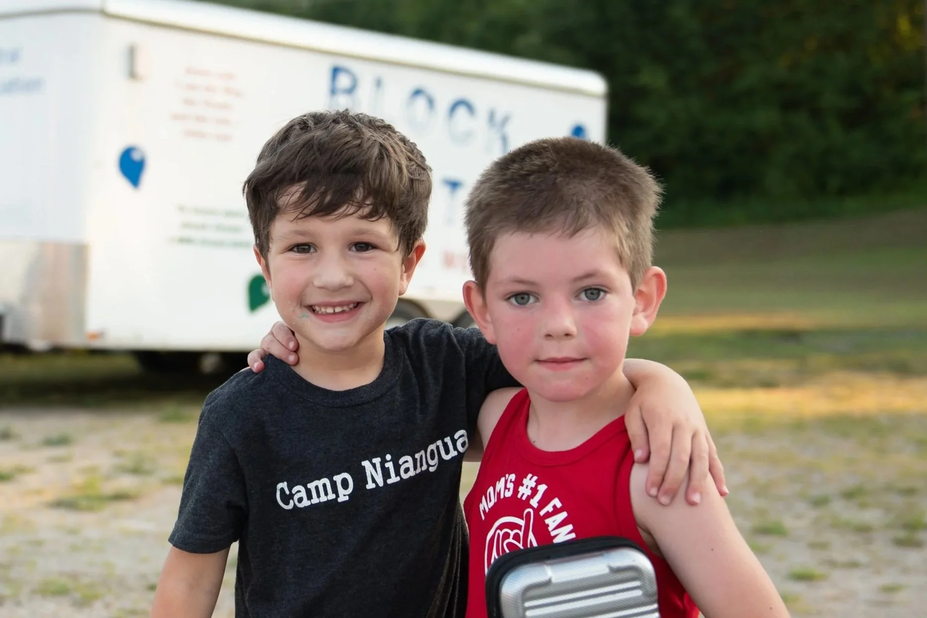 Two young boys, one with dark brown hair wearing a black t-shirt and the other with light brown hair wearing a red tank top, standing outdoors with a white trailer in the background. They are smiling and have their arms around each other.