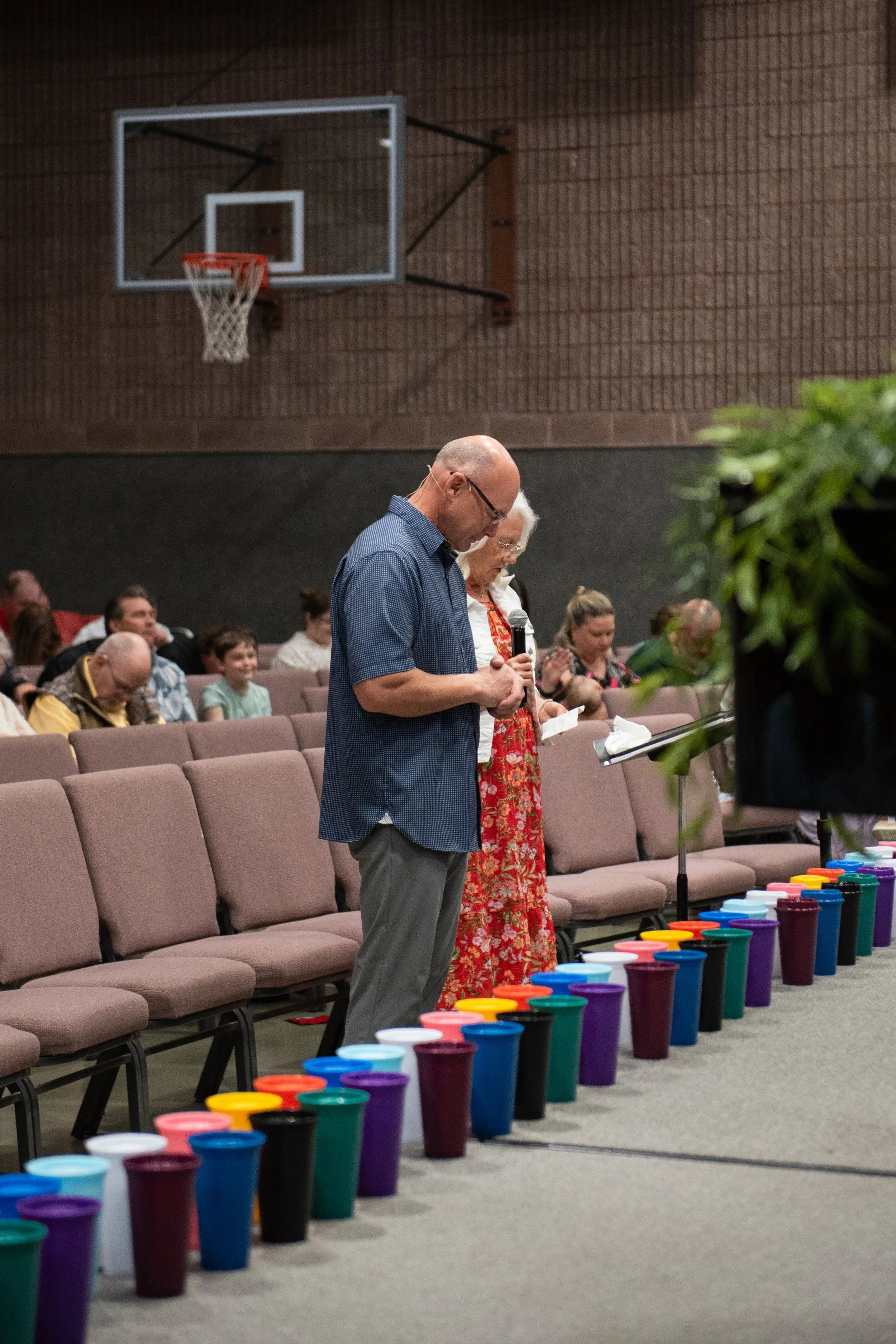 A church or community gathering with people standing and praying at the front. A row of colorful cups is arranged on the floor. A man and woman are reading or praying from papers, with the woman holding a microphone. The background shows people seated in rows in a gymnasium-style setting with a basketball hoop on the wall.