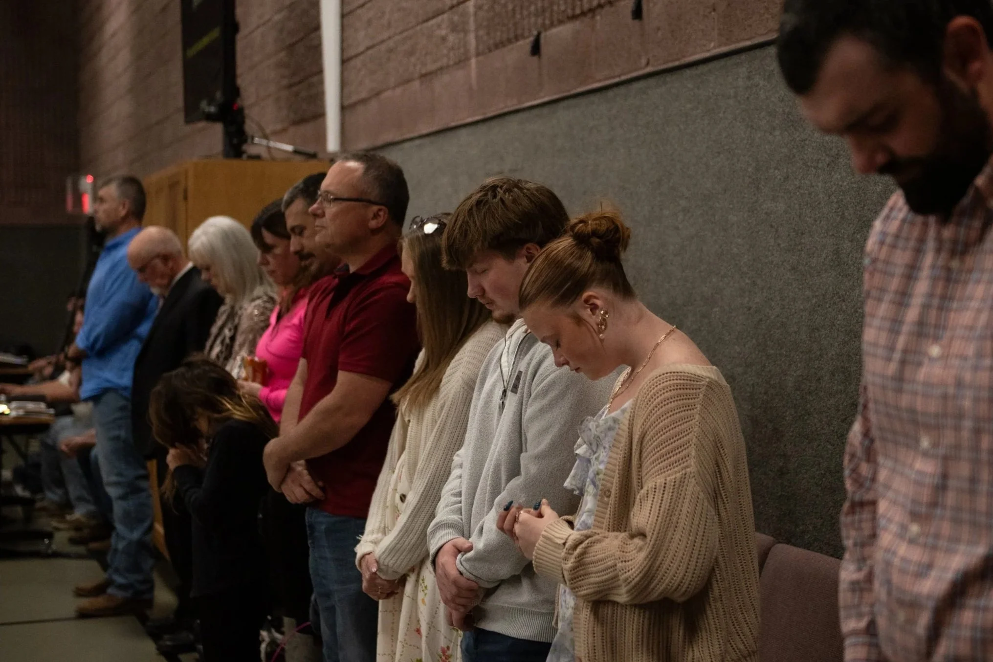 A group of people standing with heads bowed, eyes closed, and hands clasped, possibly praying or participating in a moment of silence, inside a room with a gray cushioned wall.