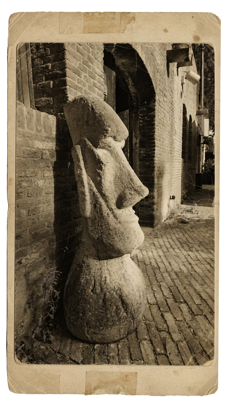 A large stone sculpture of a human face with prominent features, including a long nose and closed eyes, leaning against a brick wall in an outdoor setting with cobblestone paving.