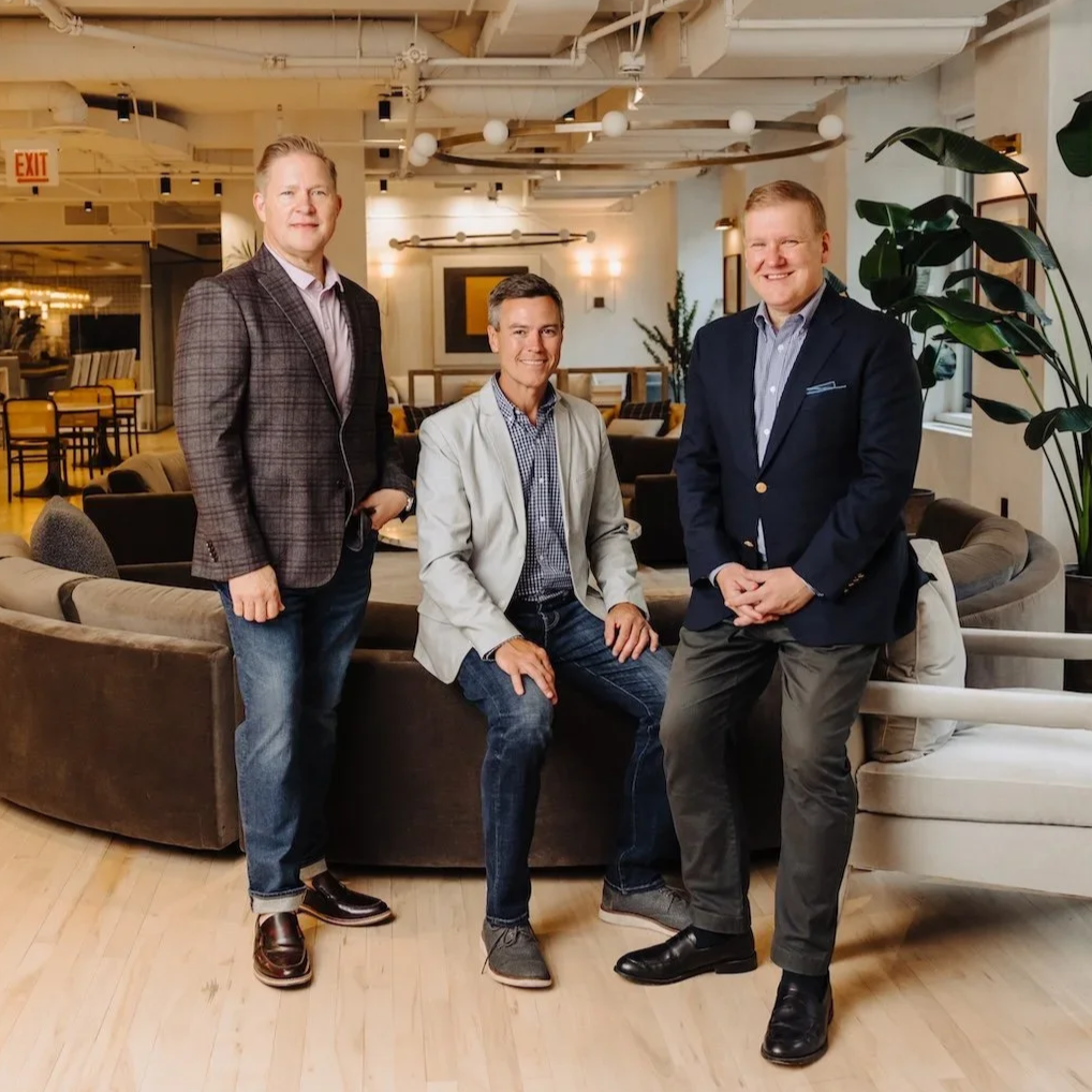 Three men in business casual attire posing together in a modern, well-lit indoor lounge area with couches, plants, and decor.
