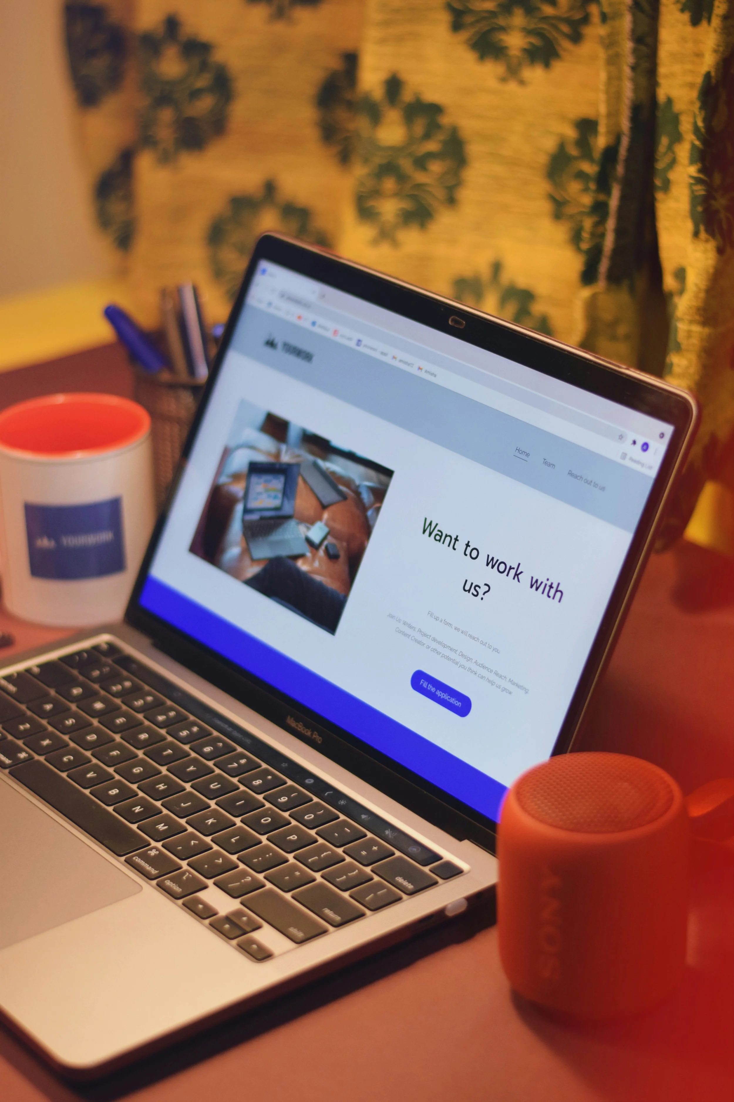 A laptop on a desk displaying a webpage for a company offering work opportunities, with an orange portable speaker and a mug nearby, and a blurred background of a curtain with leaf patterns.