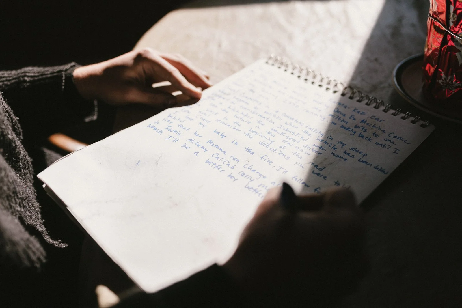 A person writing in a spiral notebook with blue ink, sitting at a table with a BPA bag on a black plate nearby.