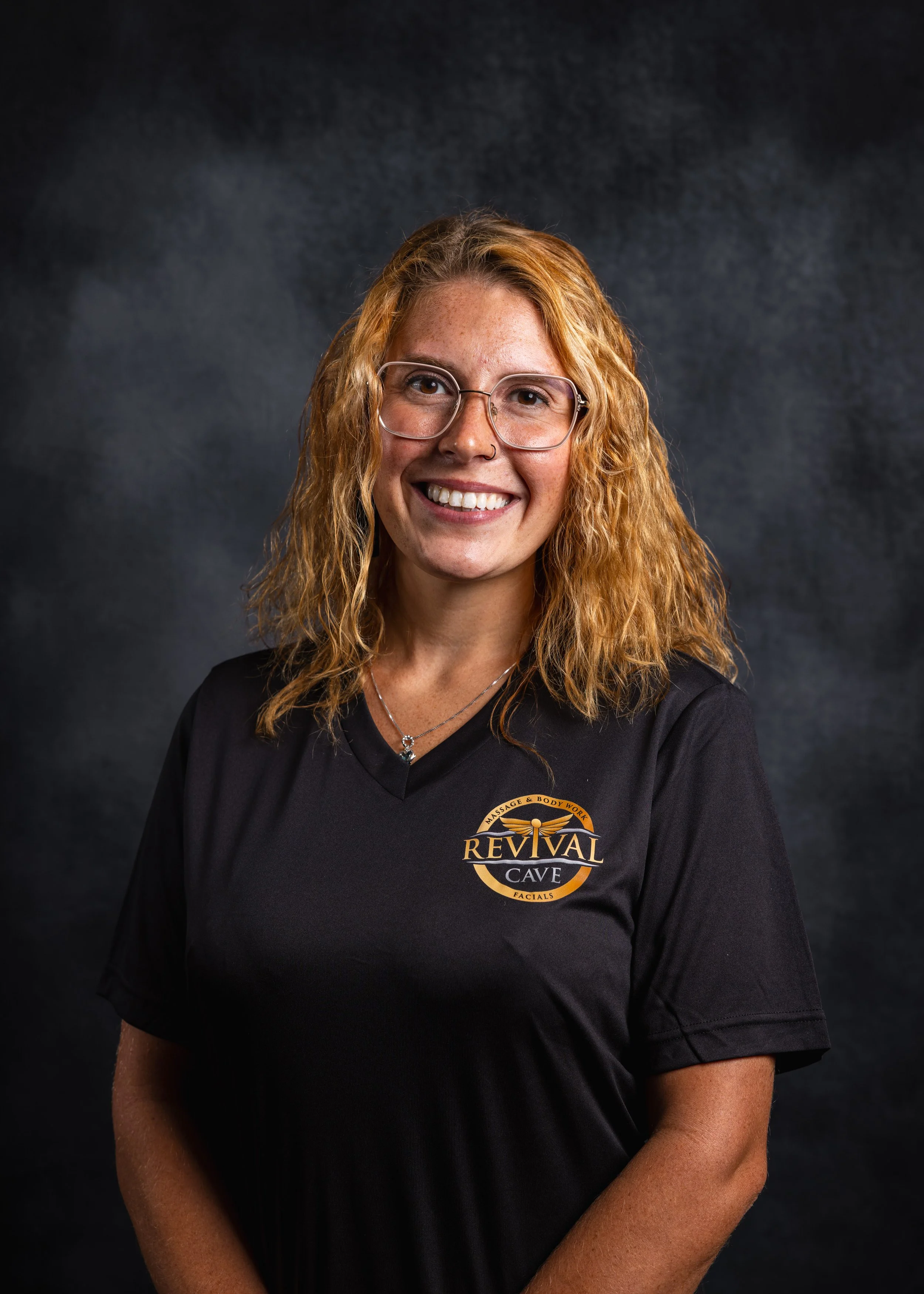 A woman with curly red hair, glasses, and a nose ring smiling at the camera. She is wearing a black shirt with a logo that reads 'Revival Cave Facials'.