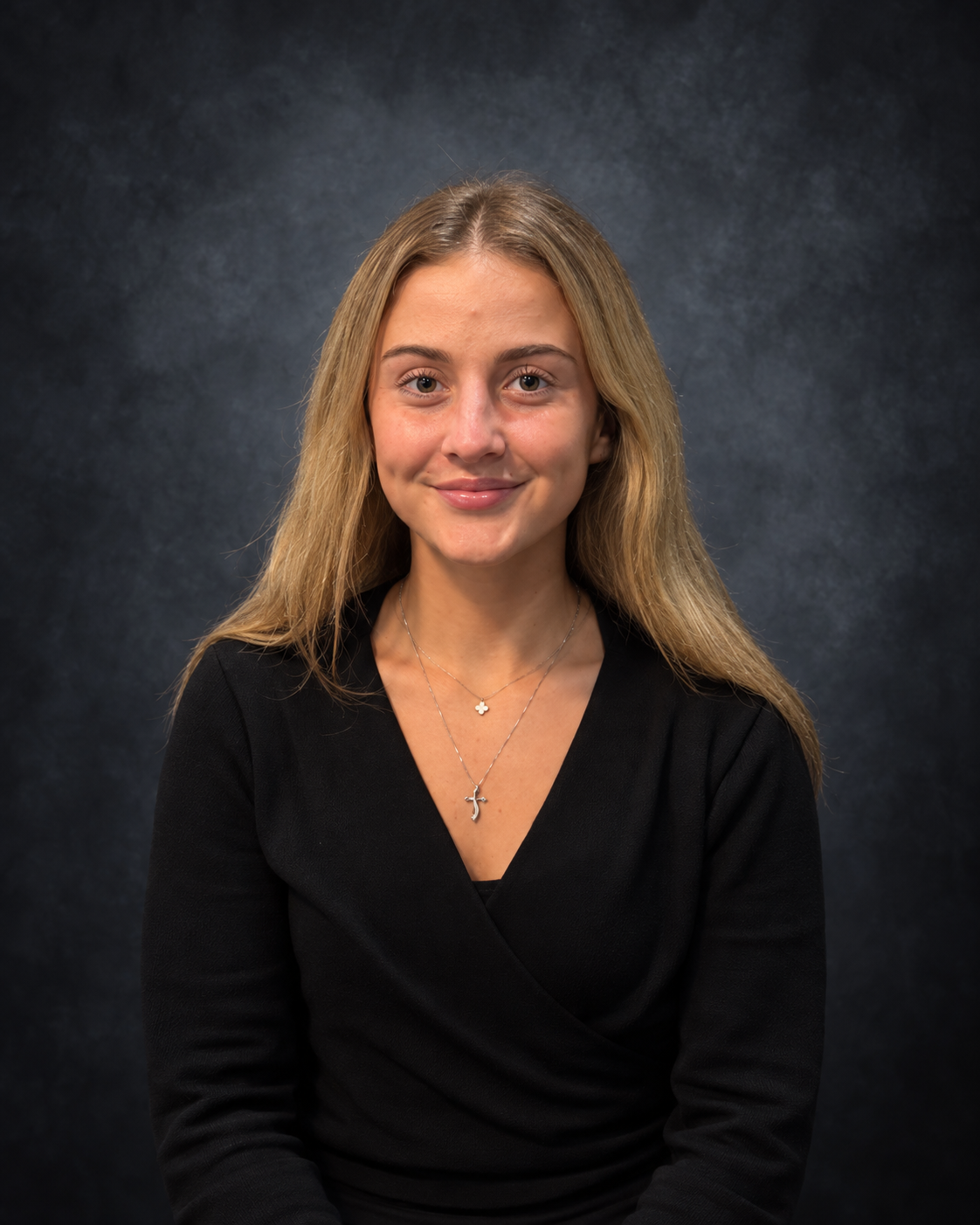 Portrait of a young woman with blonde hair, wearing a black top and necklaces, smiling at the camera against a dark background.
