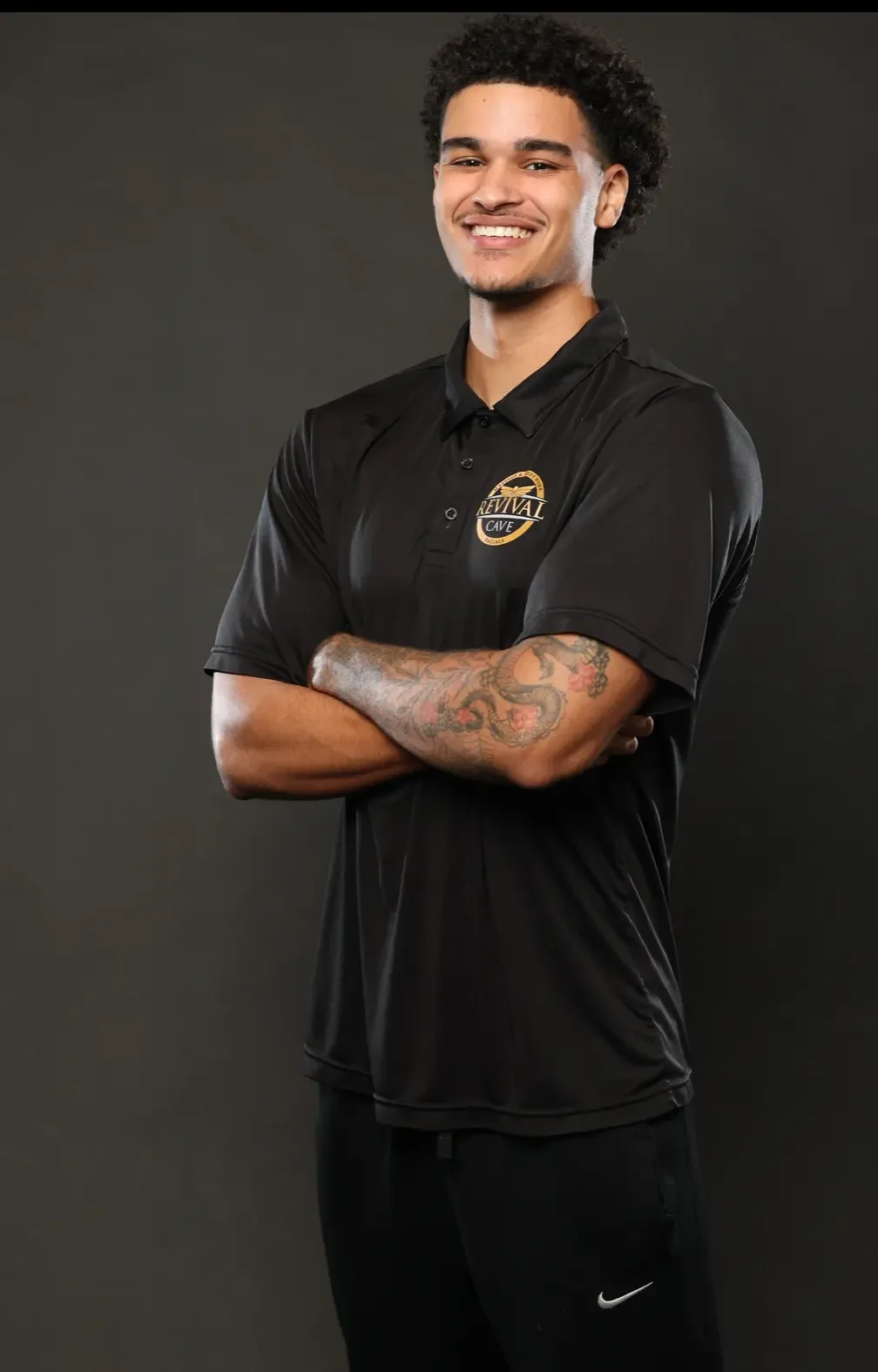 A young man with curly hair and tattoos on his arms, smiling and crossing his arms. He is wearing a black collared shirt with a logo that says 'Revival Cafe' and black Nike pants, standing against a dark background.