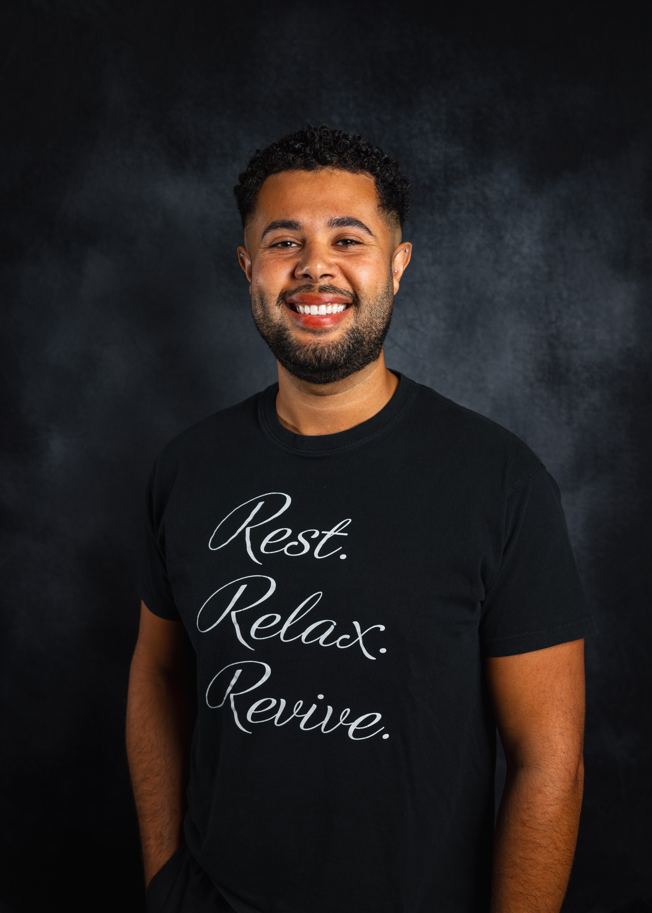 Portrait of a smiling man with a beard and curly hair, wearing a black t-shirt with the words 'Rest. Relax. Rejoice.' written on it, standing against a dark background.