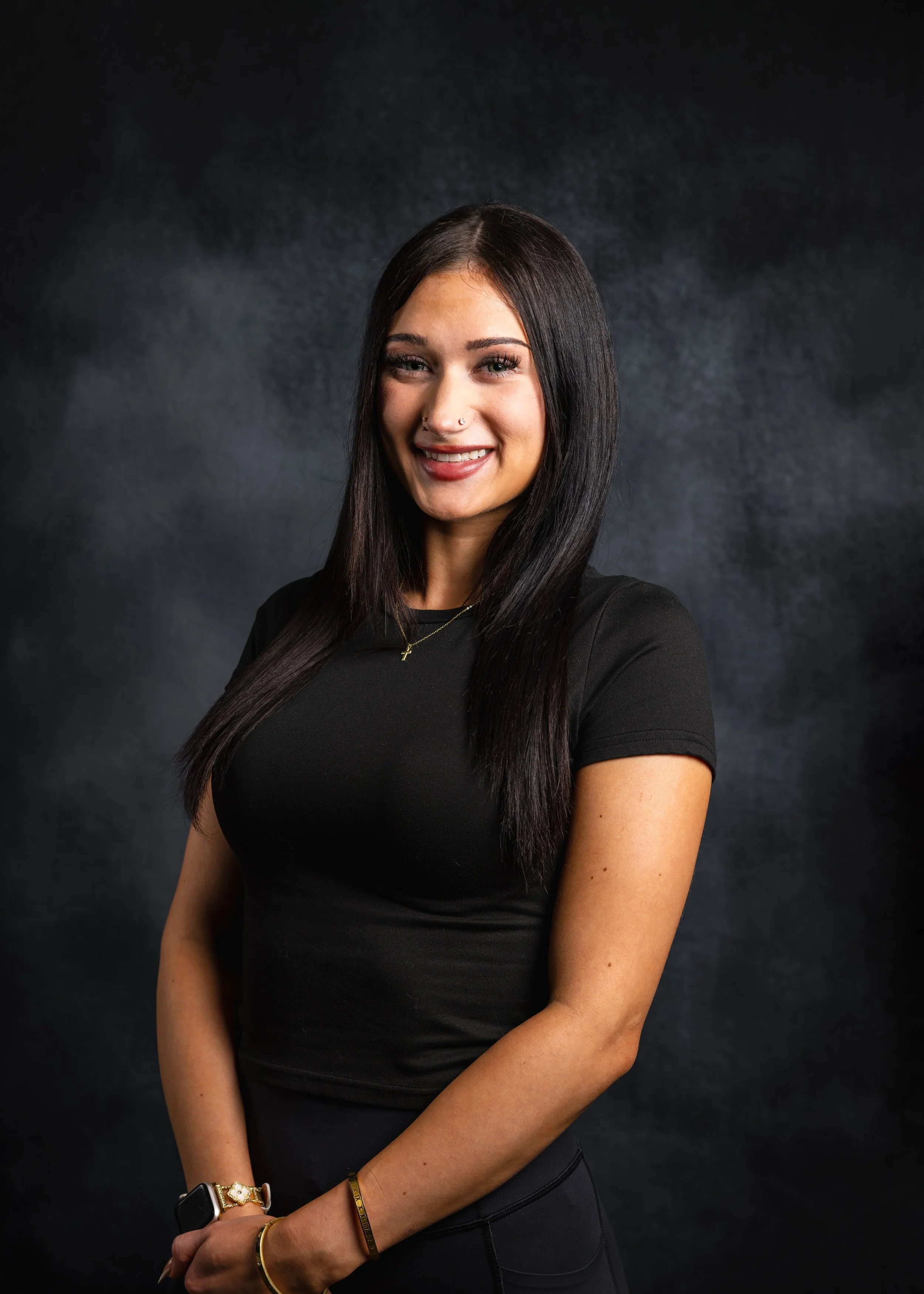 A woman with long dark hair, wearing a black shirt, jewelry, and a smartwatch, smiling in front of a dark background.