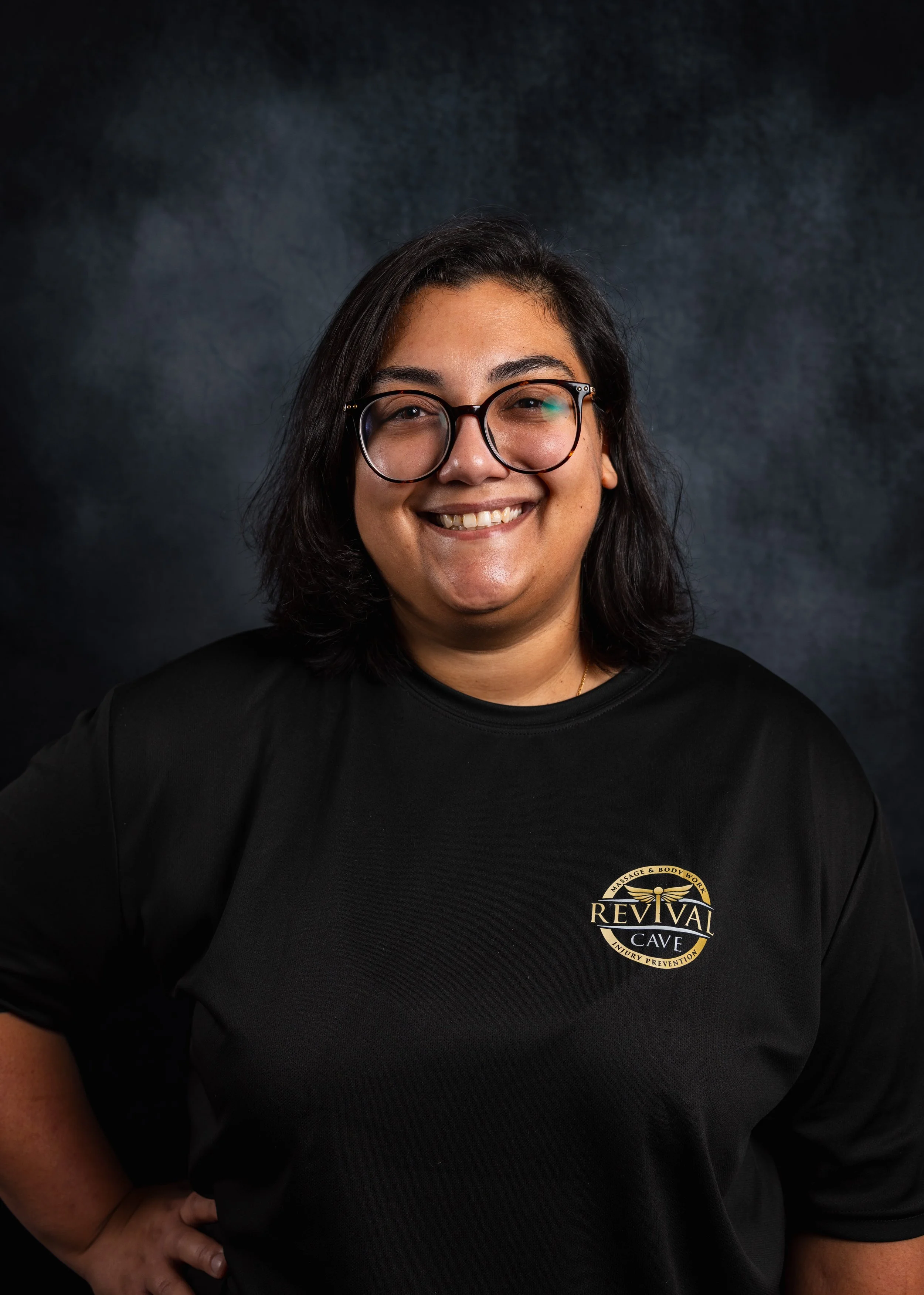 A woman with medium-length dark hair and glasses smiling, wearing a black shirt with a logo for Reviva Cave on it, standing against a dark background.