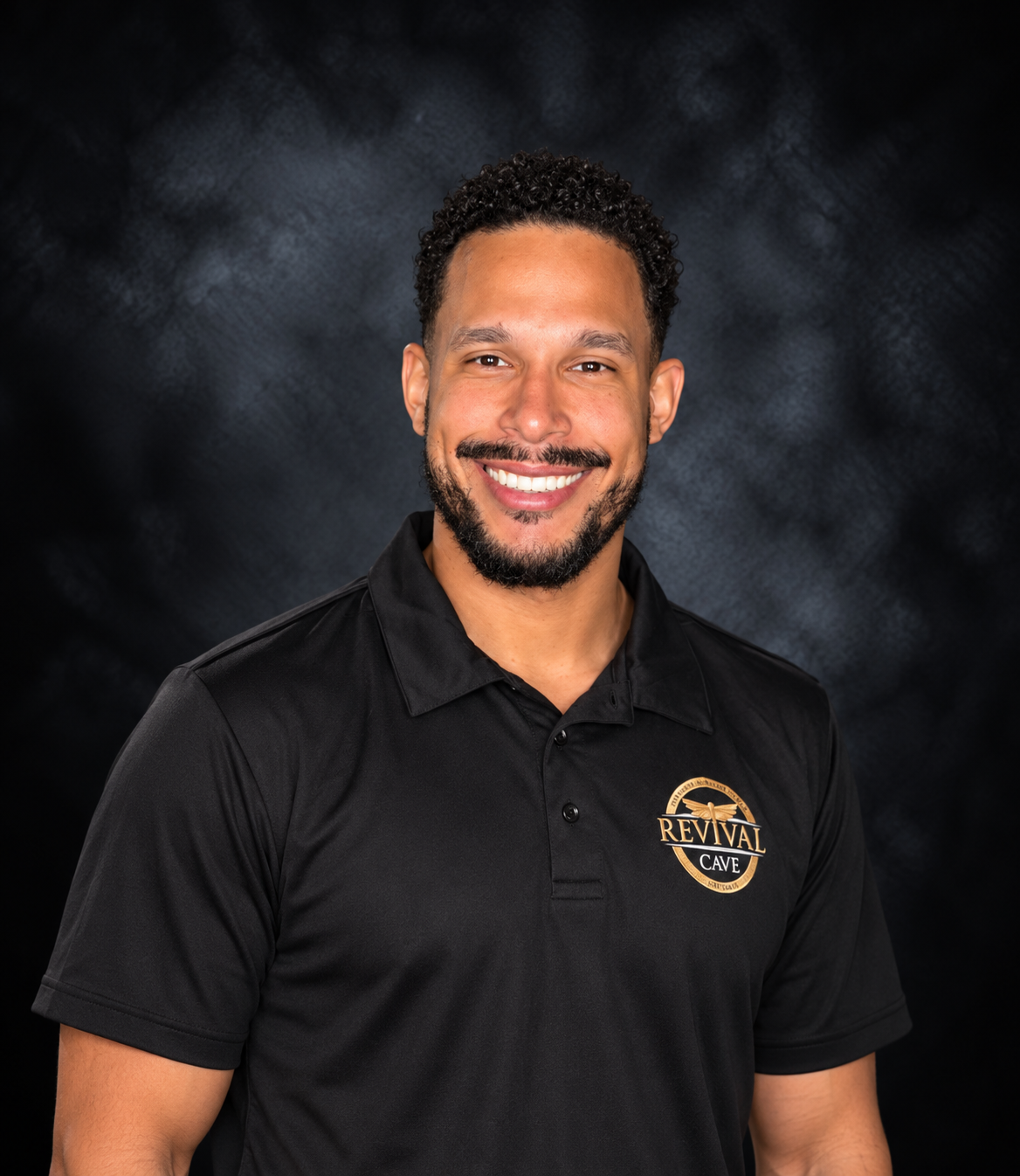 A man with short curly hair and a beard, smiling, wearing a black collared shirt with a 'Revival Cave' logo, standing against a dark background.