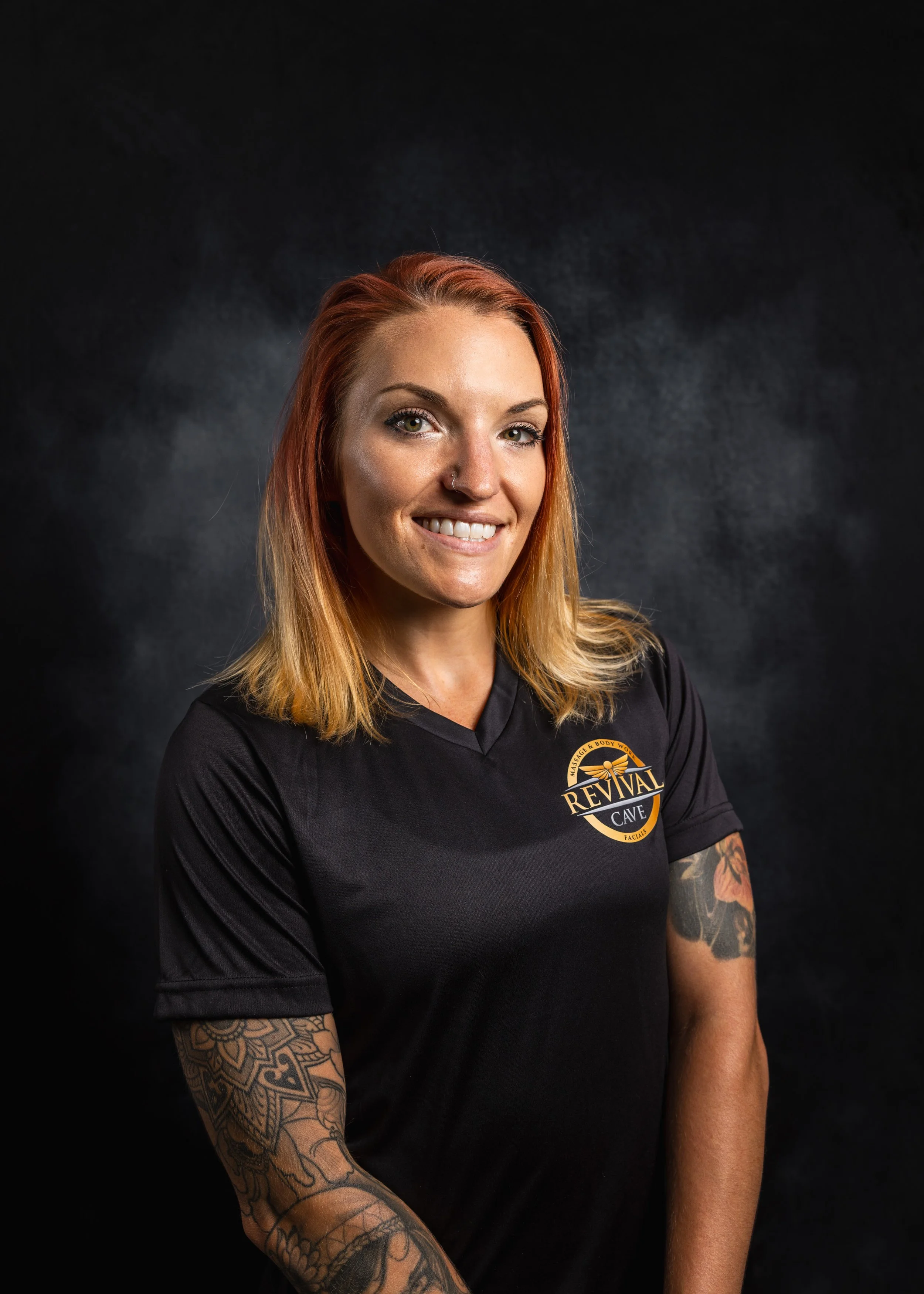 Portrait of a smiling woman with red hair, tattoos on her left arm, wearing a black shirt with a logo that says 'Revival Cave' on it, against a dark background.
