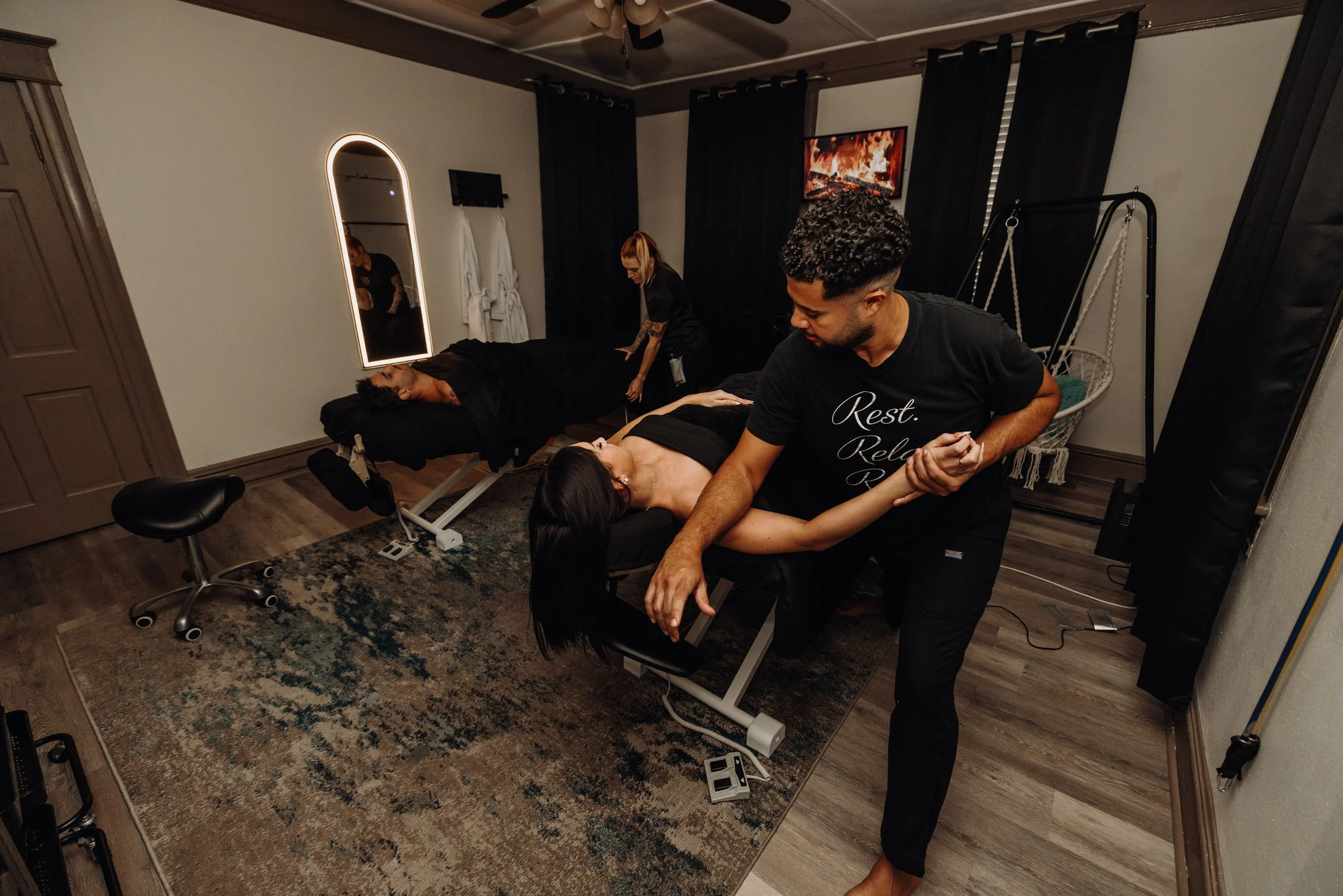A person is lying face up on a massage table, receiving a massage from another person while a third person stands nearby. The room has a television on the wall showing fire, a mirror, and a swing chair in the corner.