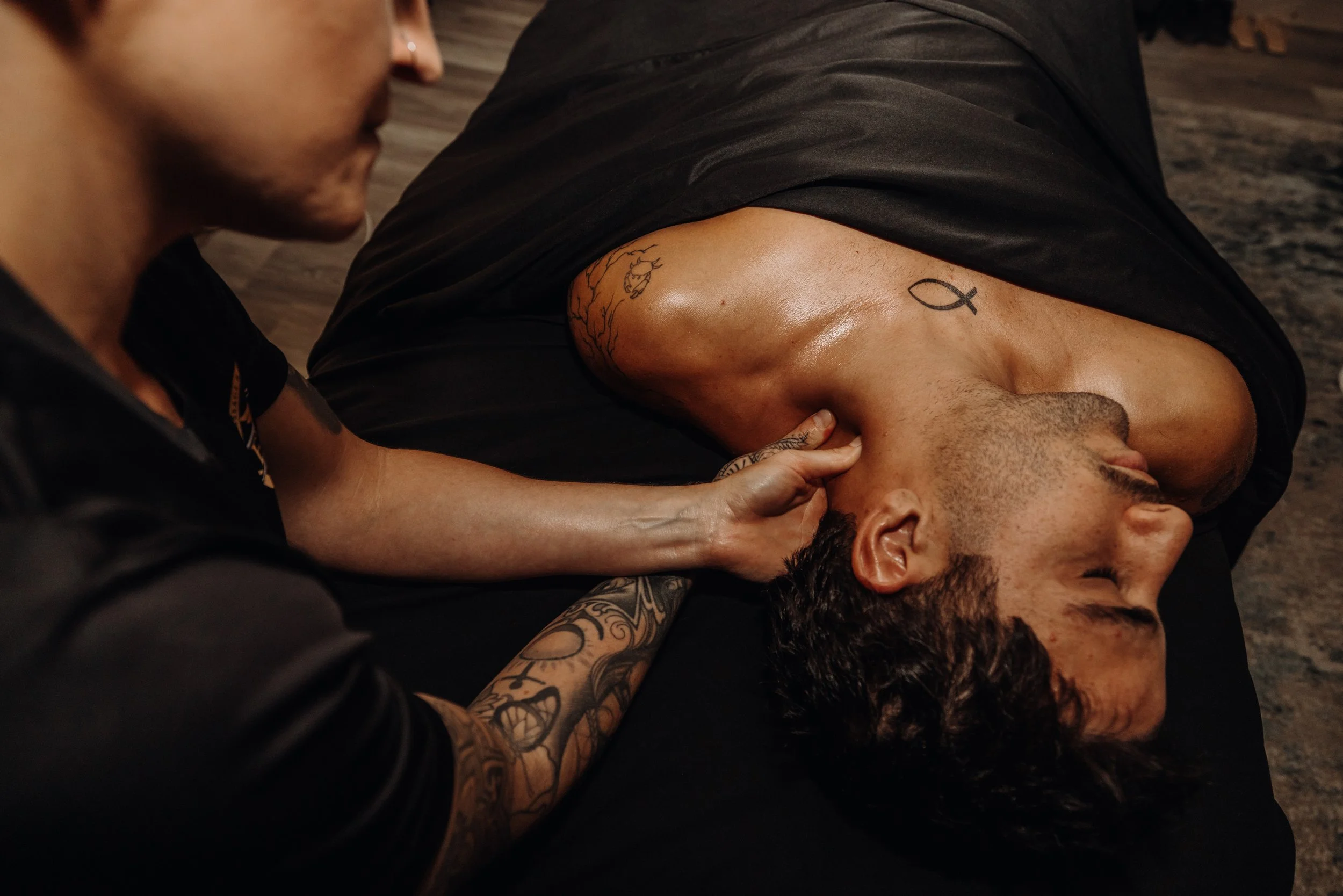 A tattooed woman in black clothing holds a man who is lying down with his eyes closed, tattoos visible on his arm and neck, in a dimly lit room with a dark floor.