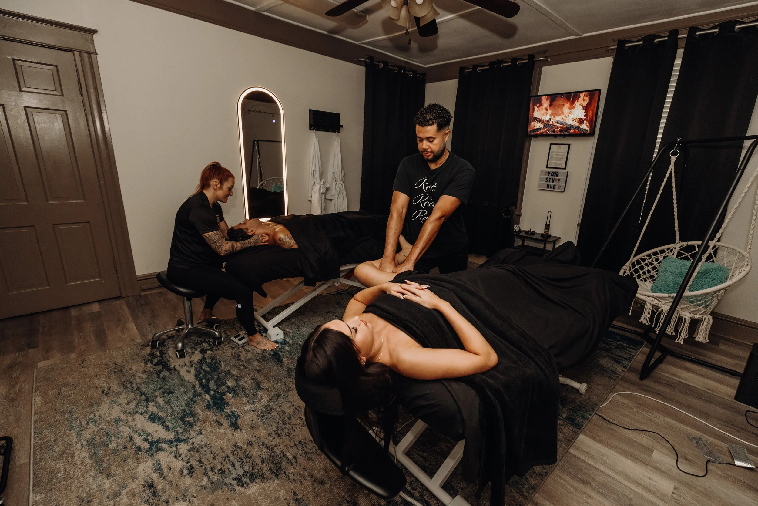A massage therapist performs a massage on a woman lying face-up on a massage table, with another woman receiving a massage in the foreground. A television shows a fireplace, and a decorative swing hangs nearby in a cozy, dimly lit room.