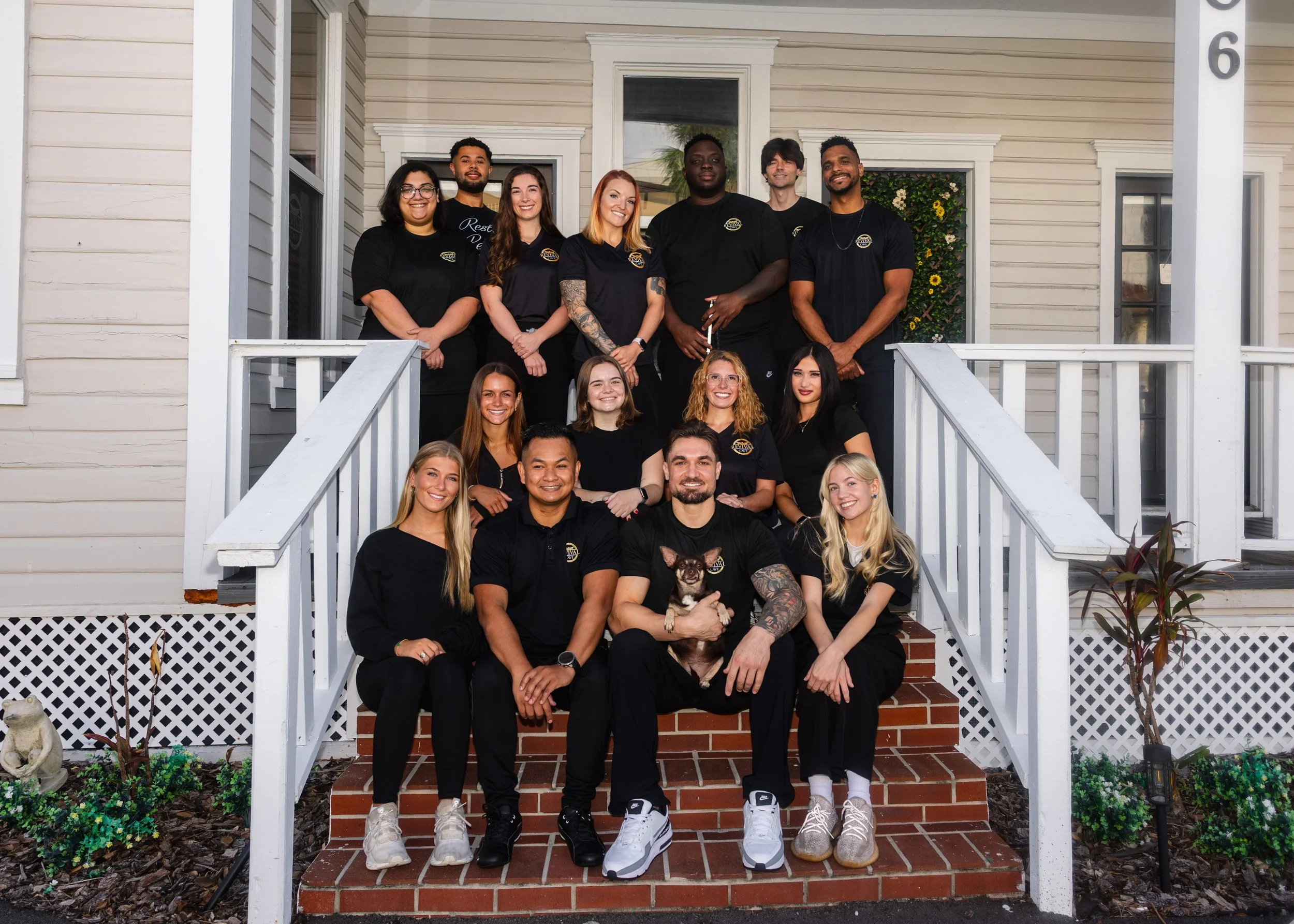 A group of sixteen teenagers and young adults, along with a small dog, gathered on the front steps of a house, posing for a photo. They are smiling and dressed in casual clothes, mainly black t-shirts, with some wearing glasses, tattoos, and various hairstyles.