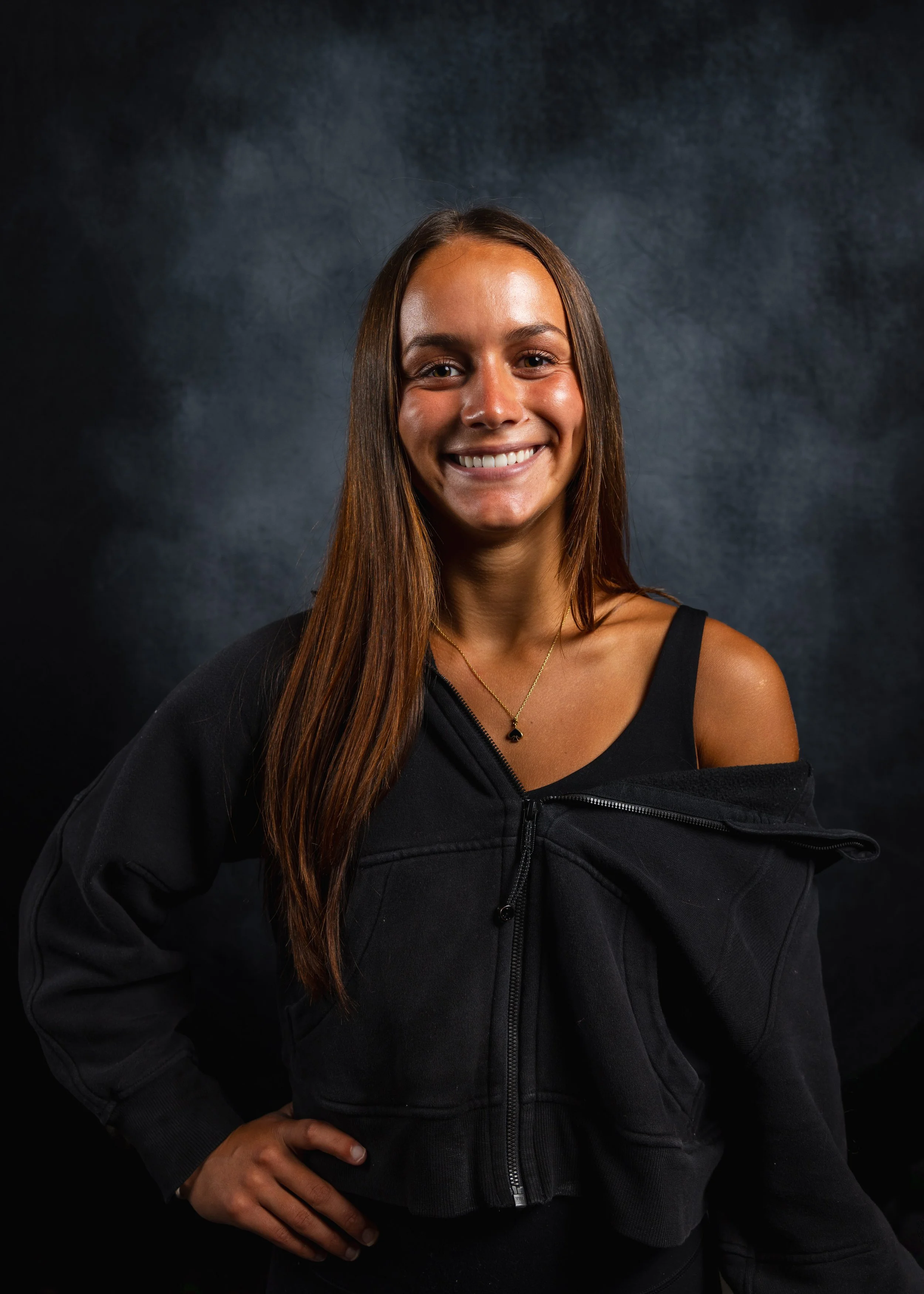 A young woman with long brown hair, smiling, wearing a black zip-up jacket over a black top and a gold necklace, standing against a dark background.