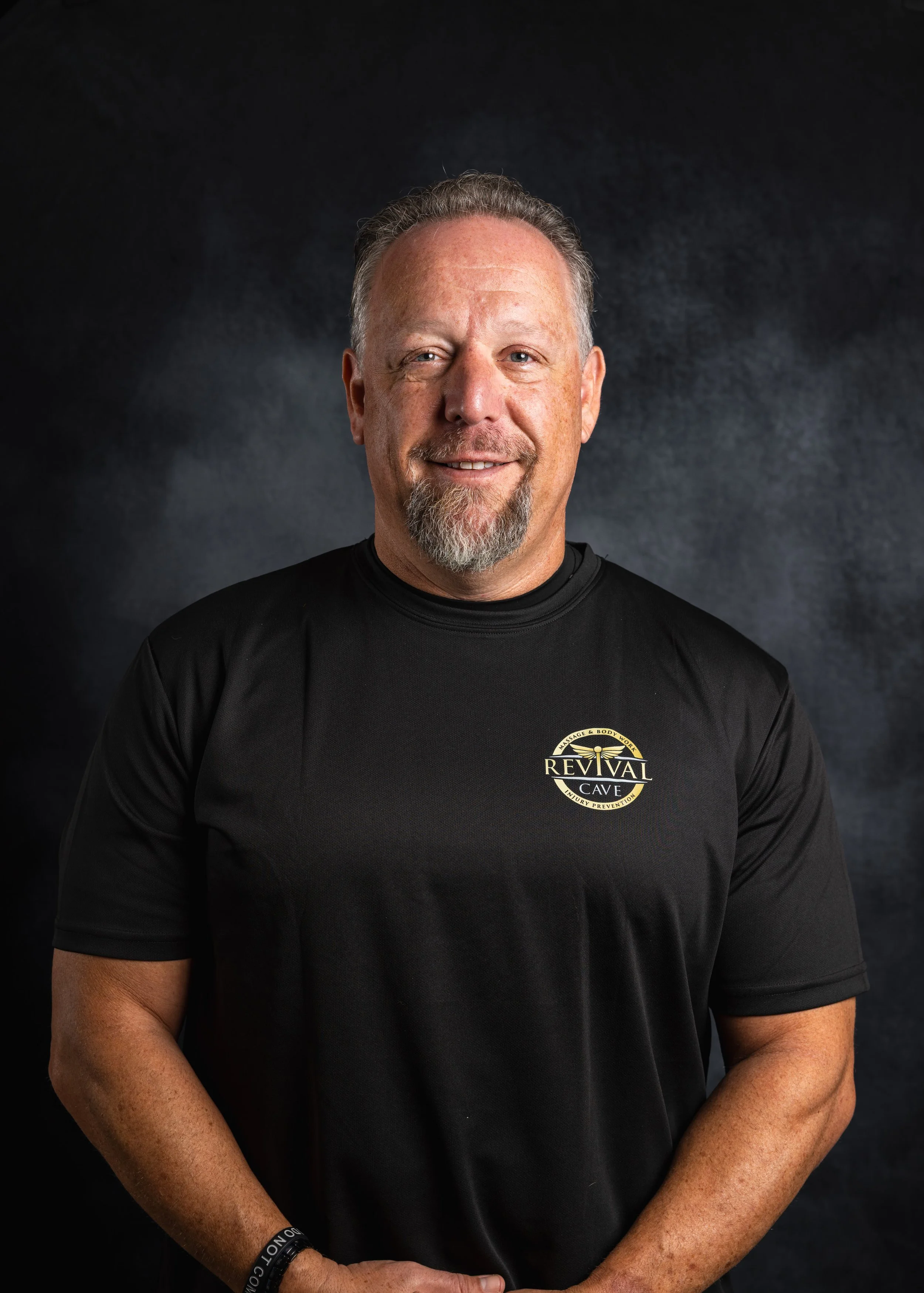 A middle-aged man with short, gray hair and a beard smiling at the camera, wearing a black t-shirt with a logo that says 'Revival Cave' on the front, against a dark, textured background.