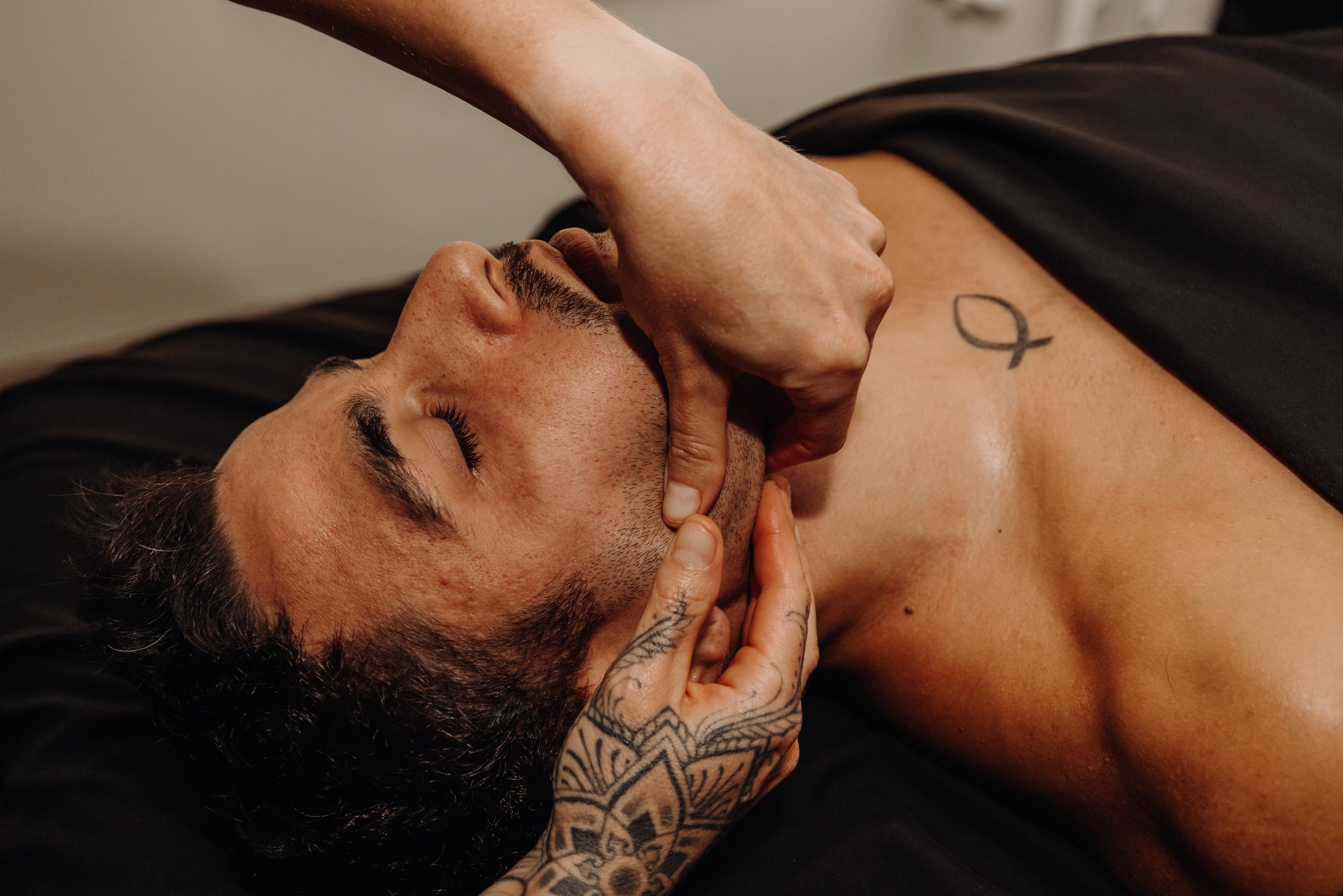A man with tattoos on his arm and chest lying down with his eyes closed while a person touches his face during a massage or therapy session.