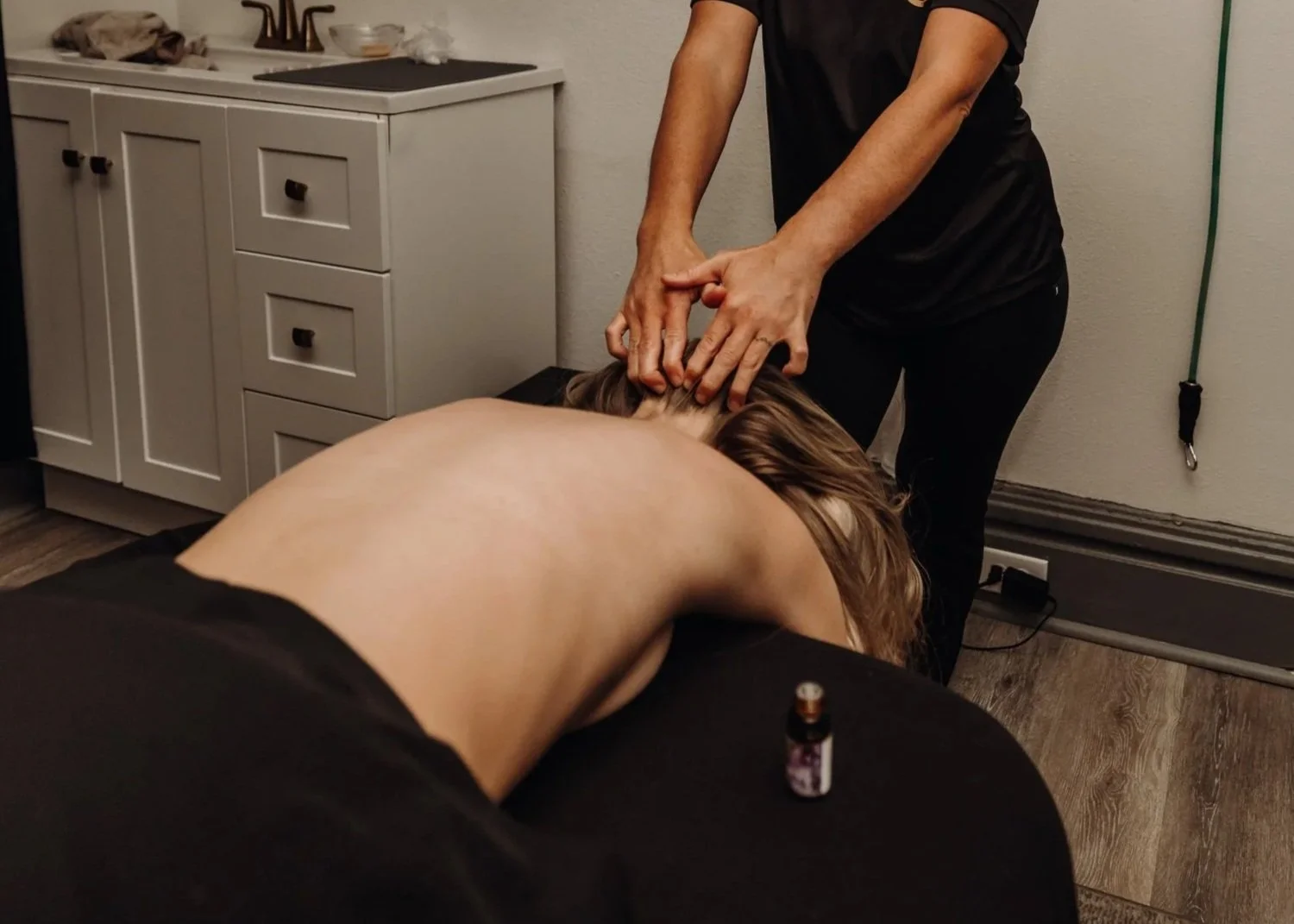 A person receiving a massage or acupuncture, lying face down on a massage table, with a practitioner applying pressure to their neck. A small bottle of essential oil is on the table nearby, and the room has a wooden floor and a white cabinet in the background.
