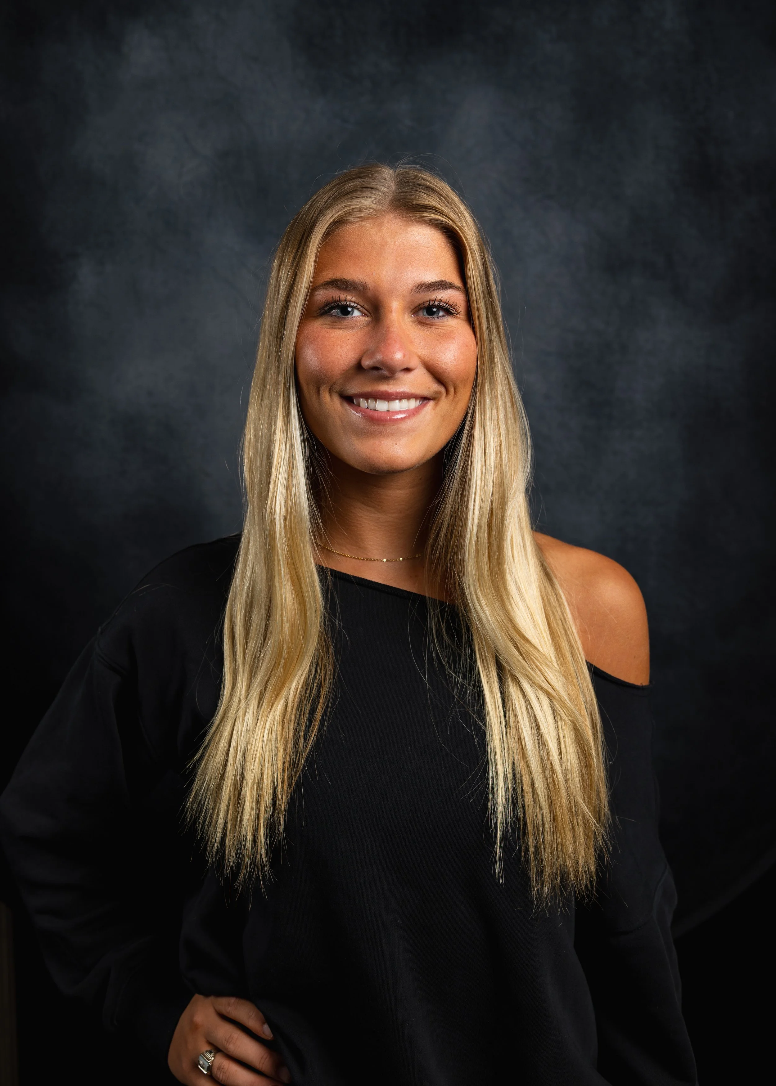 A woman with long blonde hair, blue eyes, and a bright smile, wearing a black top with shoulder cutouts, stands against a dark, textured background.