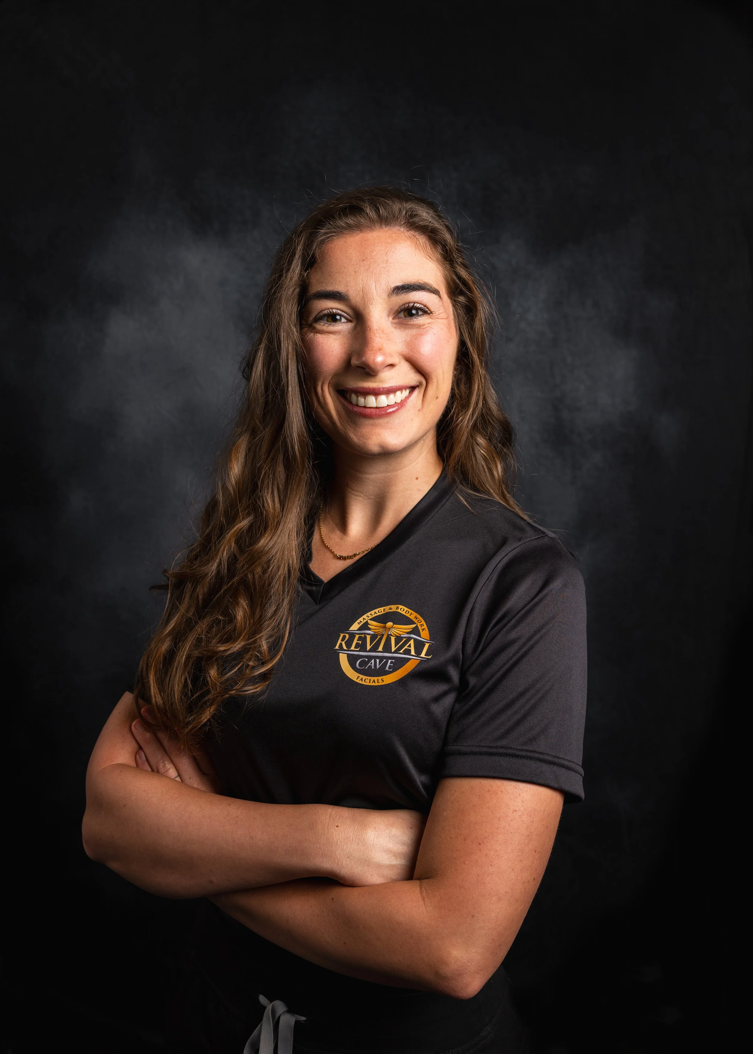 A smiling woman with long, wavy brown hair, wearing a black shirt with a logo that reads 'Revival Cave' and has her arms crossed, standing against a dark background.