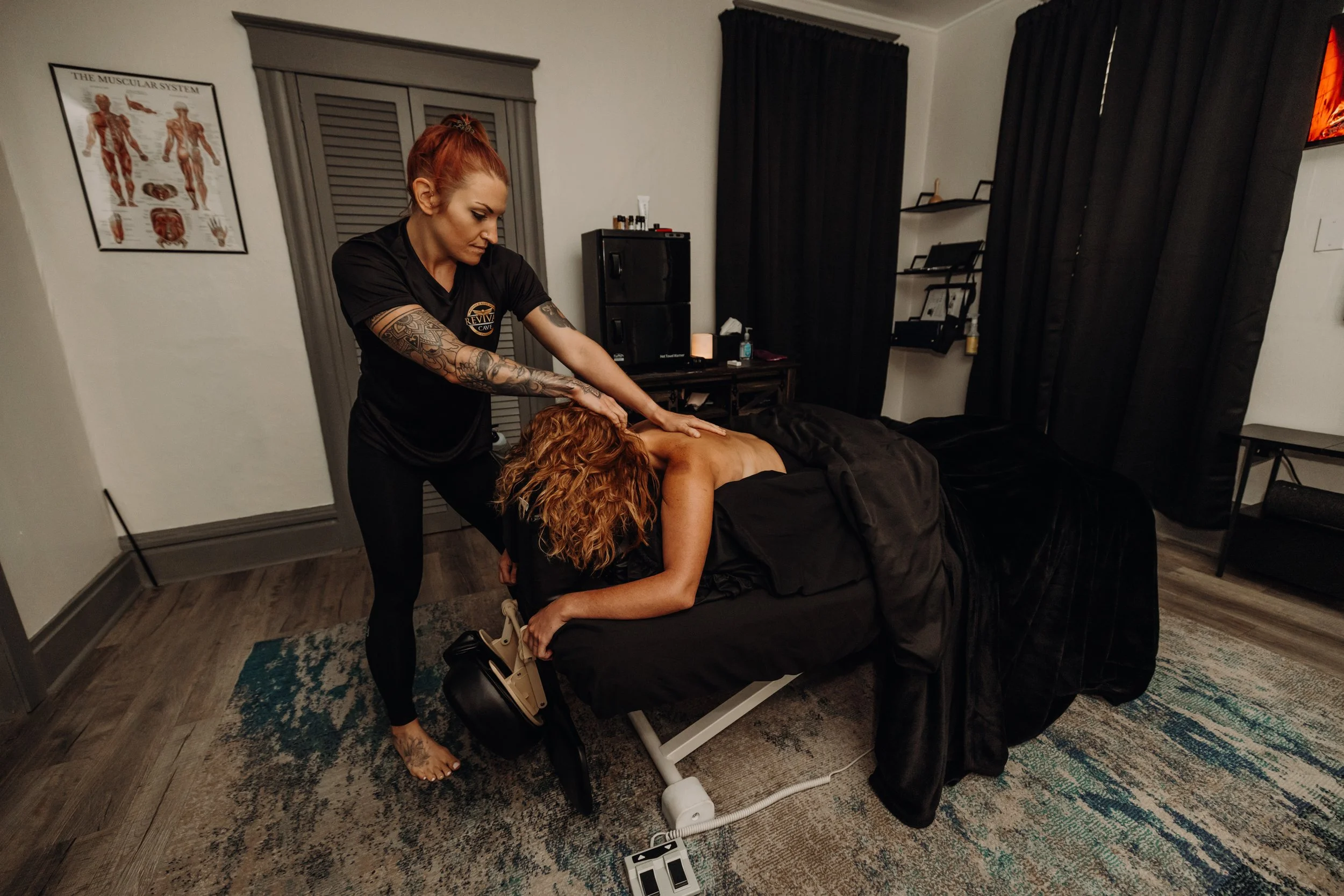 A woman receiving a back massage from a massage therapist in a treatment room with dark curtains and medical posters.